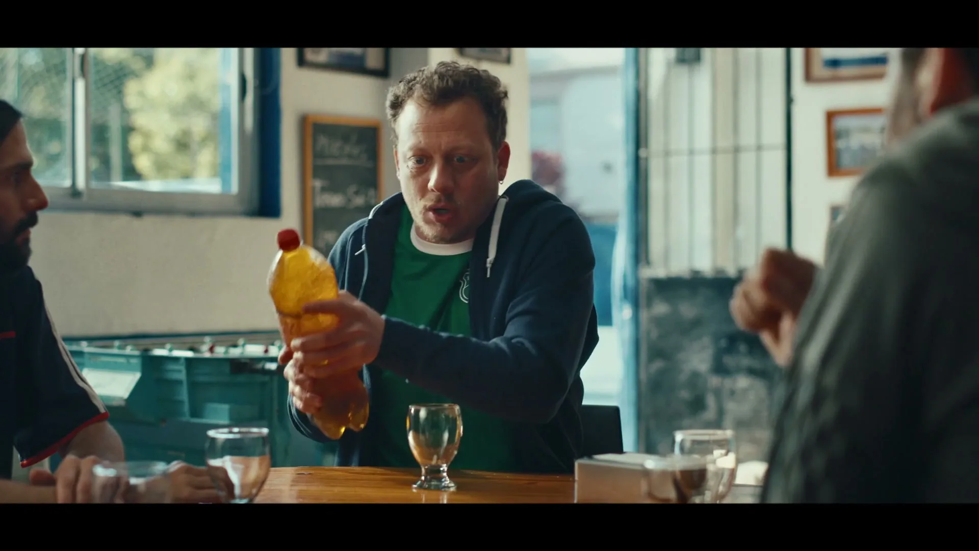 A man in his 30s, wearing a green shirt and blue hoodie, gestures with a large plastic bottle of H2oh! while talking to friends at a table in a bar.