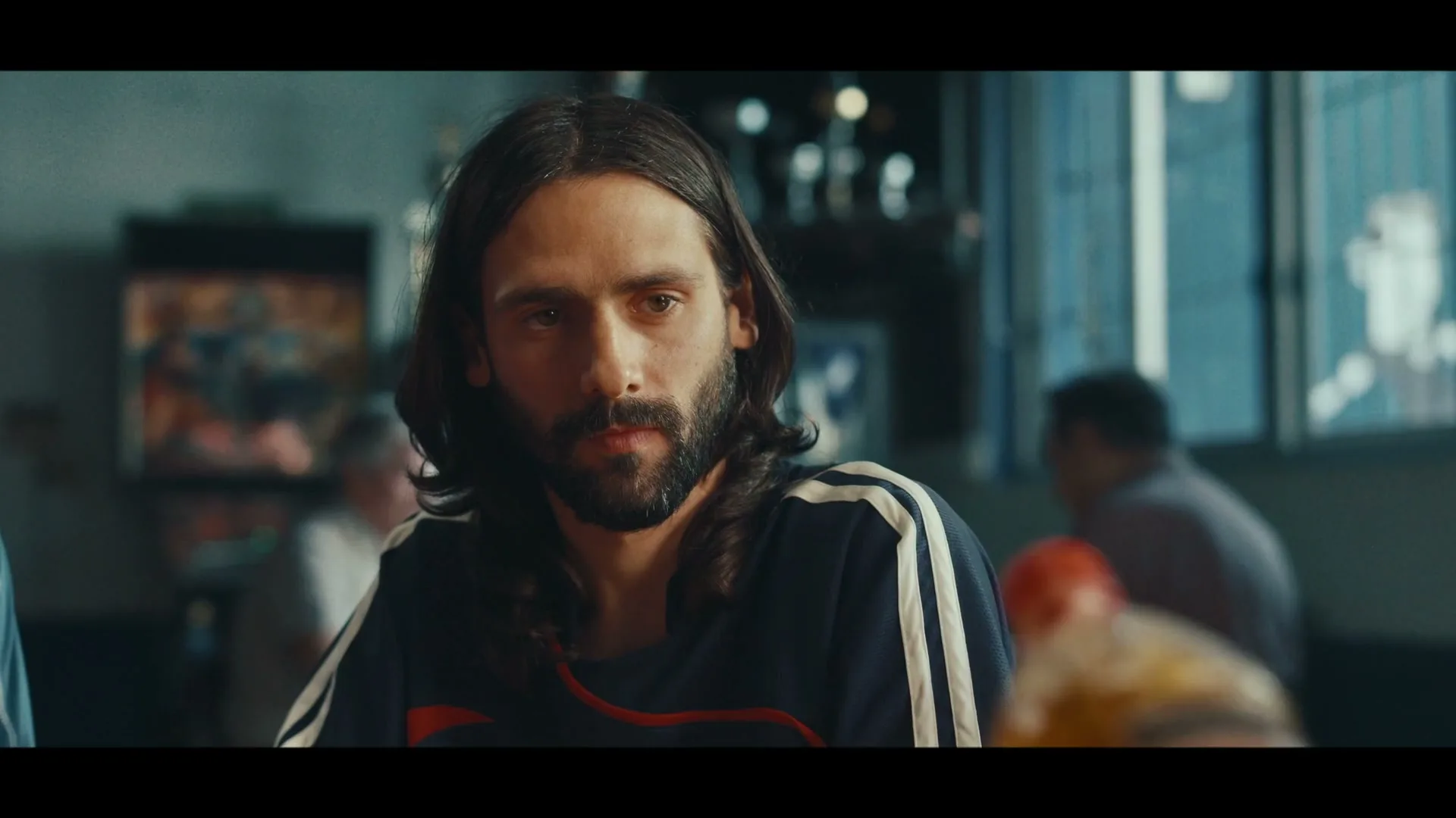 A man with long hair and a beard, wearing a dark blue shirt with white stripes, looks intently at an orange bottle being offered to him in a dimly lit bar.