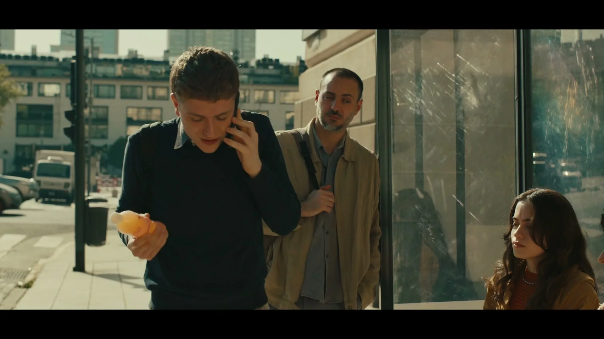 A young man in a blue sweater talks on the phone while holding an orange bottle, walking past a bus stop on a sunny city street.