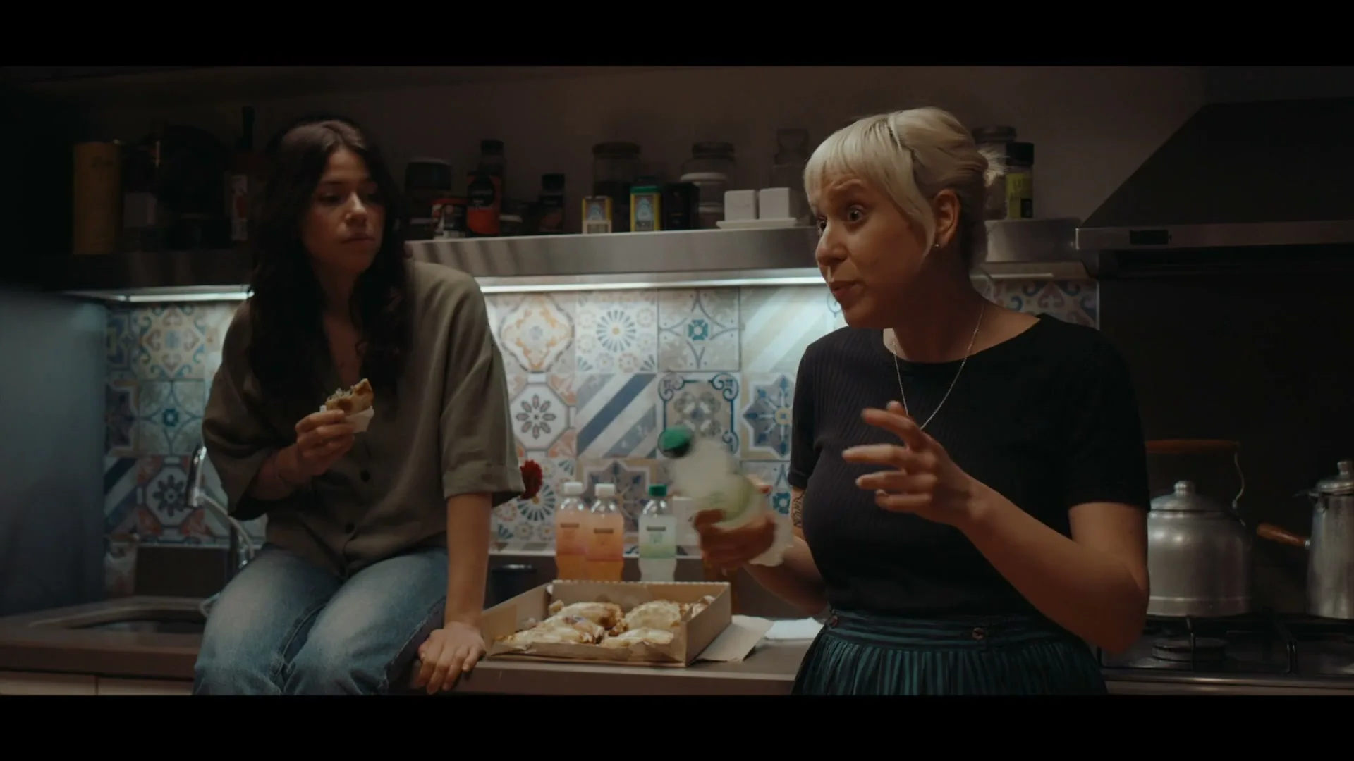 Two women are in a kitchen, one sitting on the counter eating, the other standing and talking animatedly while holding a bottle.