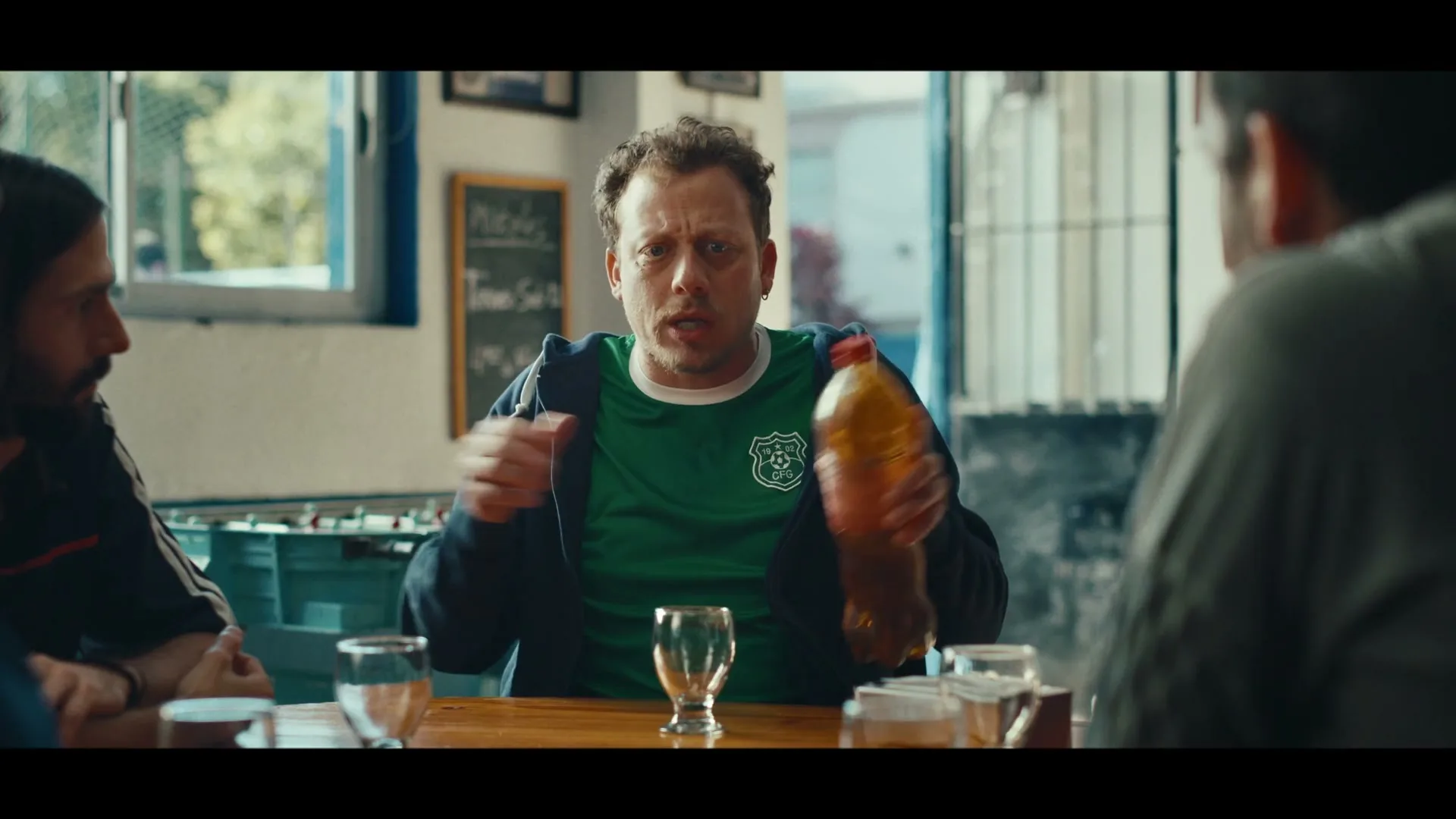 A man in his 30s, wearing a green soccer jersey and blue hoodie, sits at a table with friends, gesturing animatedly while holding a bottle of H2oh! in a casual bar setting.