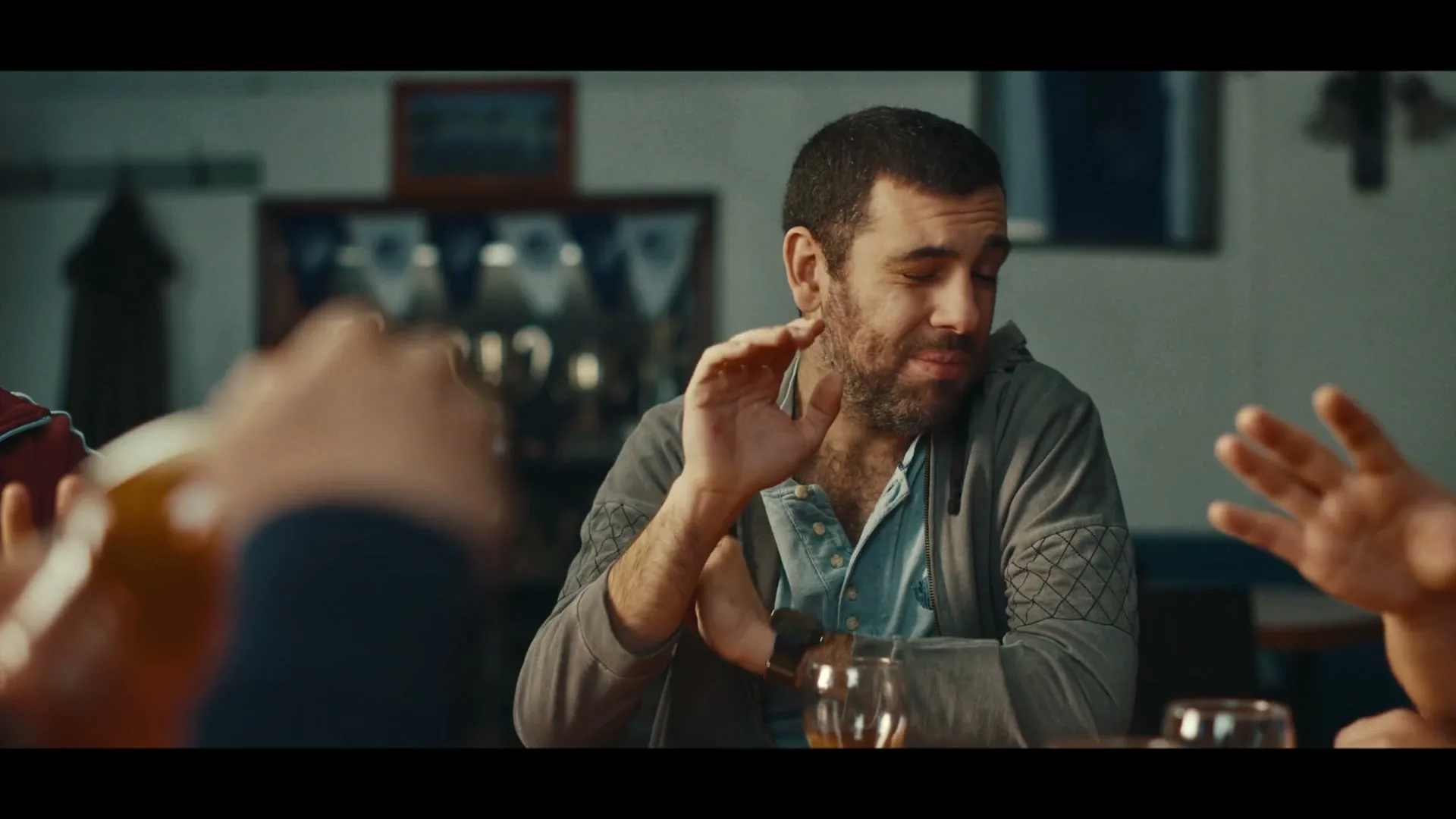 A man with a beard and a grey hoodie sits at a table, gesturing with his hands as if to say "stop" or "no" in an interior setting.
