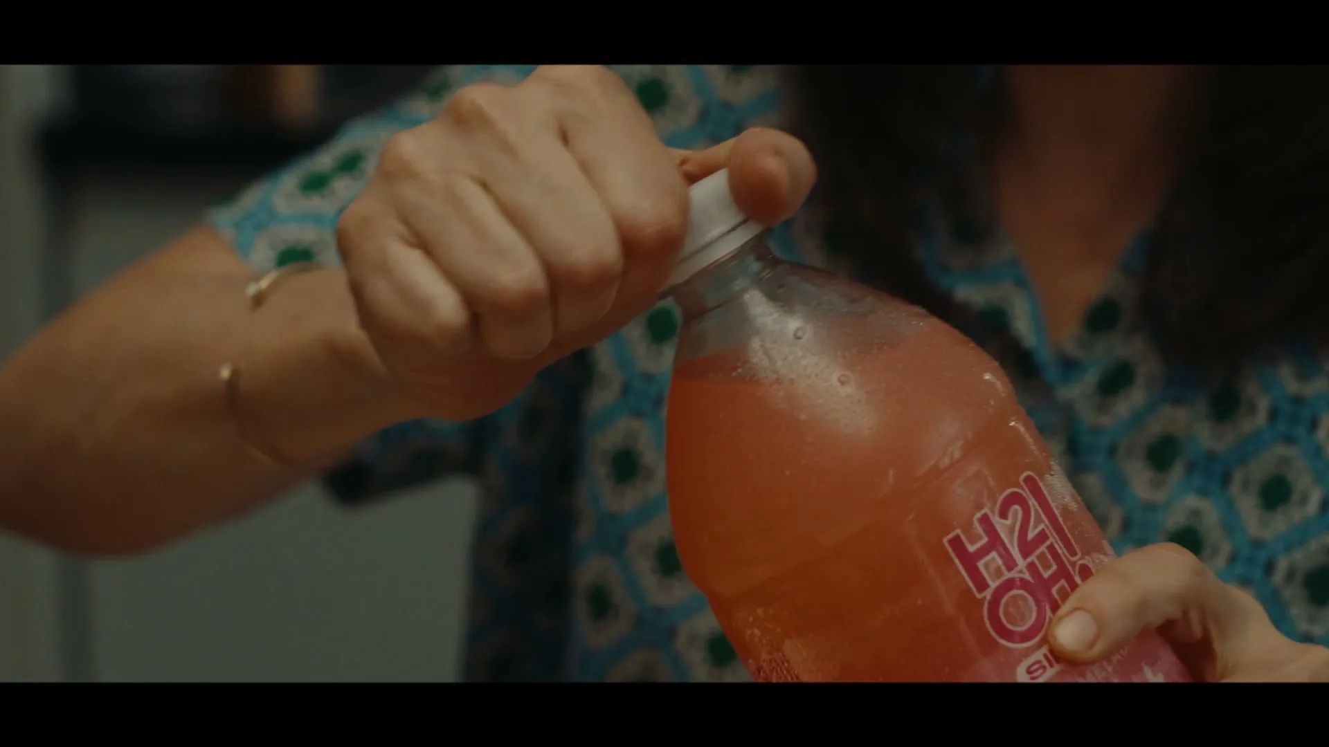 A woman's hands are shown in a medium closeup, opening a bottle of H2oh! Pomelada soda, in an interior setting.