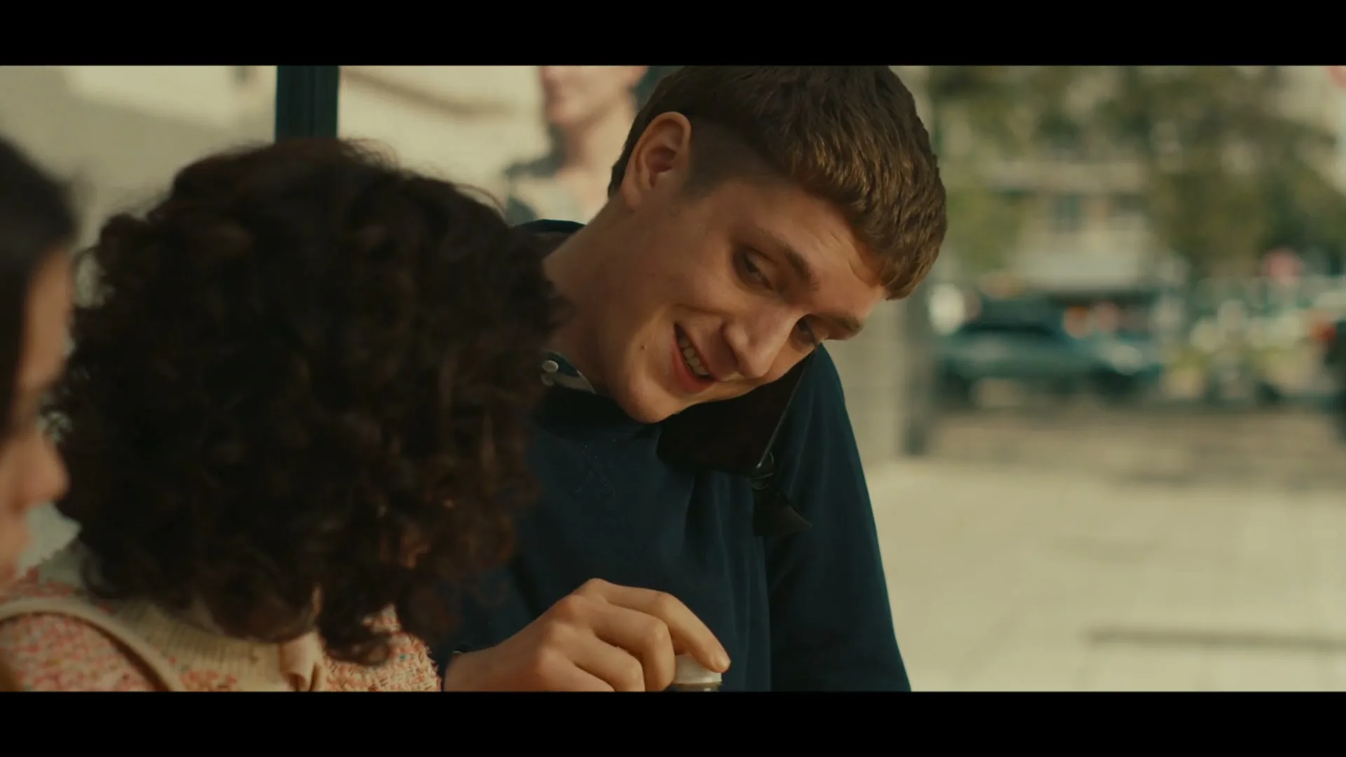 A young man with a bowl cut talks on the phone and gestures with his finger while sitting next to a woman with curly hair at an outdoor cafe.