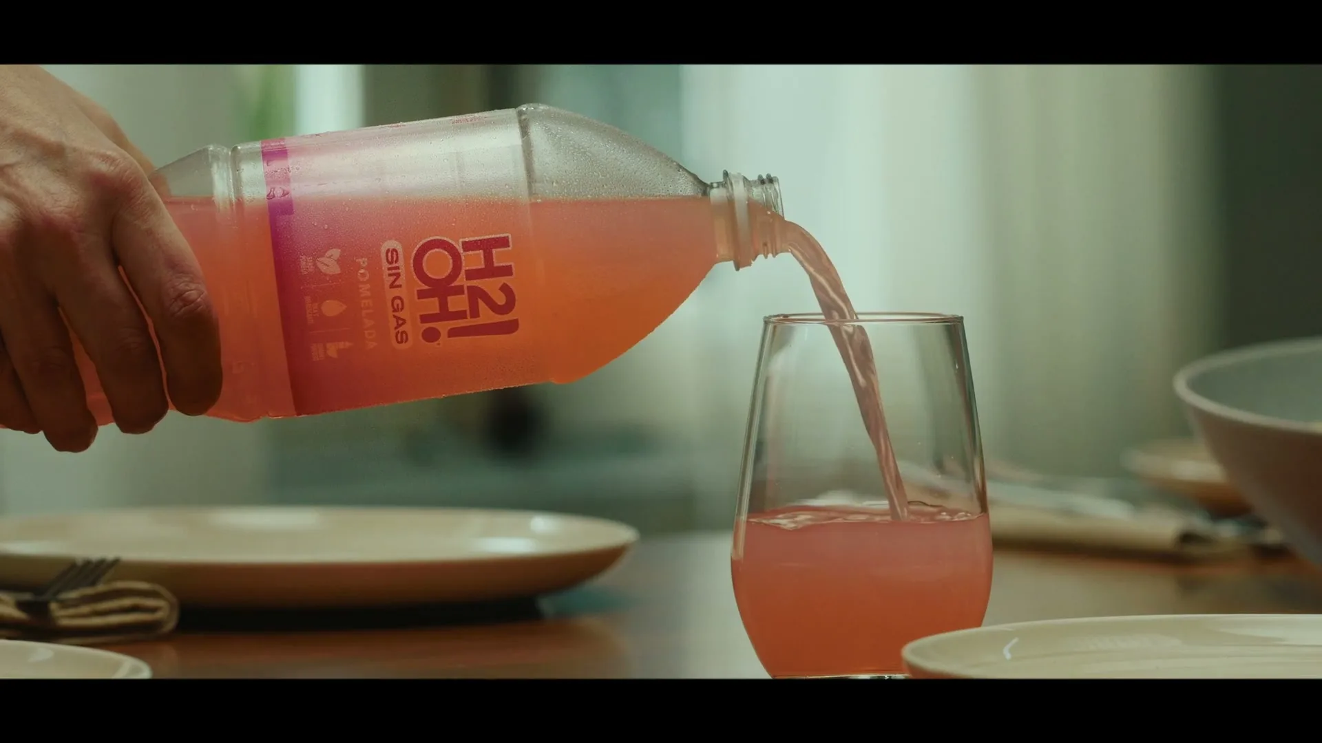 A person's hand pours pink H2oh! Pomelada drink from a plastic bottle into a glass on a wooden table set with plates.