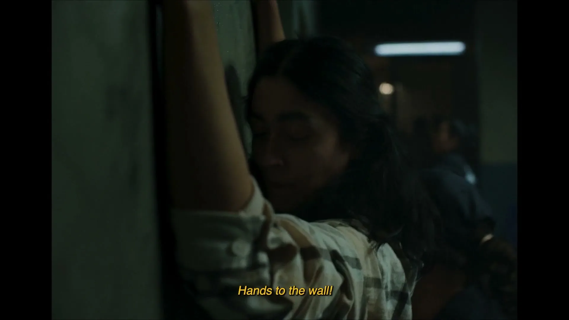A young woman with dark hair is pressed against a wall by an unseen force, looking distressed as others are pushed around her in a dark, confined space.