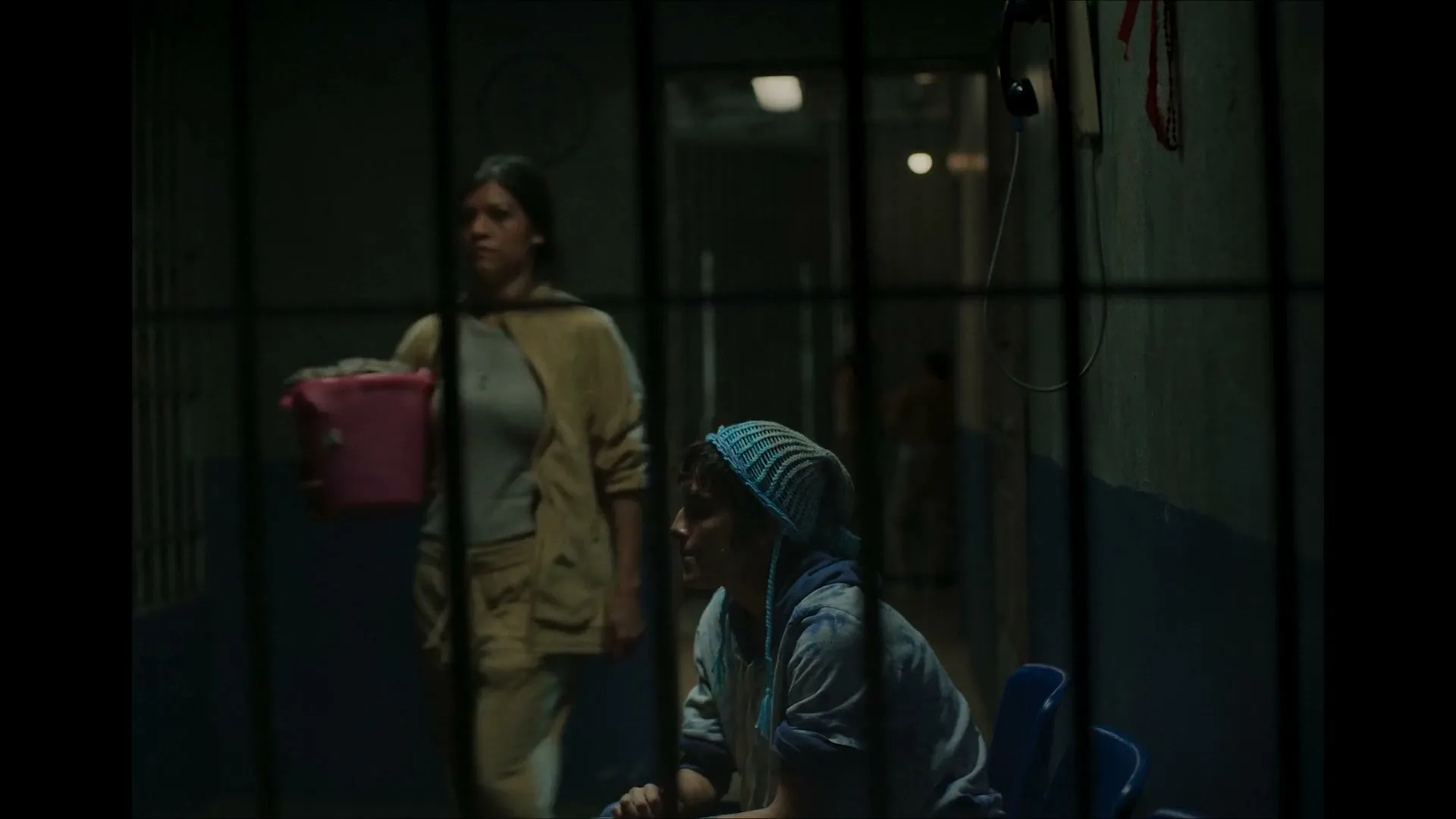A young woman in a blue headscarf sits on a chair behind prison bars, looking out as another woman walks past in a dimly lit prison hallway.