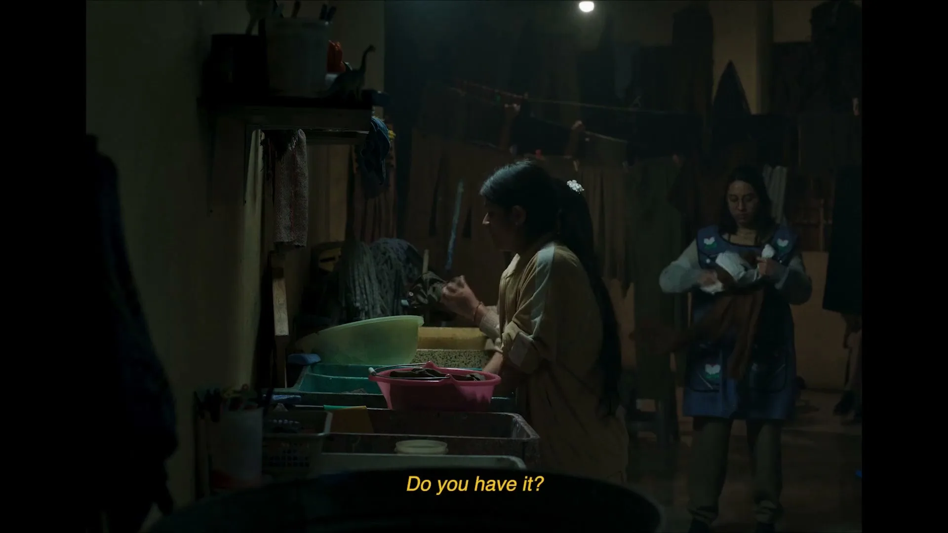Three women are in a dimly lit room, two of them washing clothes in basins, while the third hangs laundry in the background.