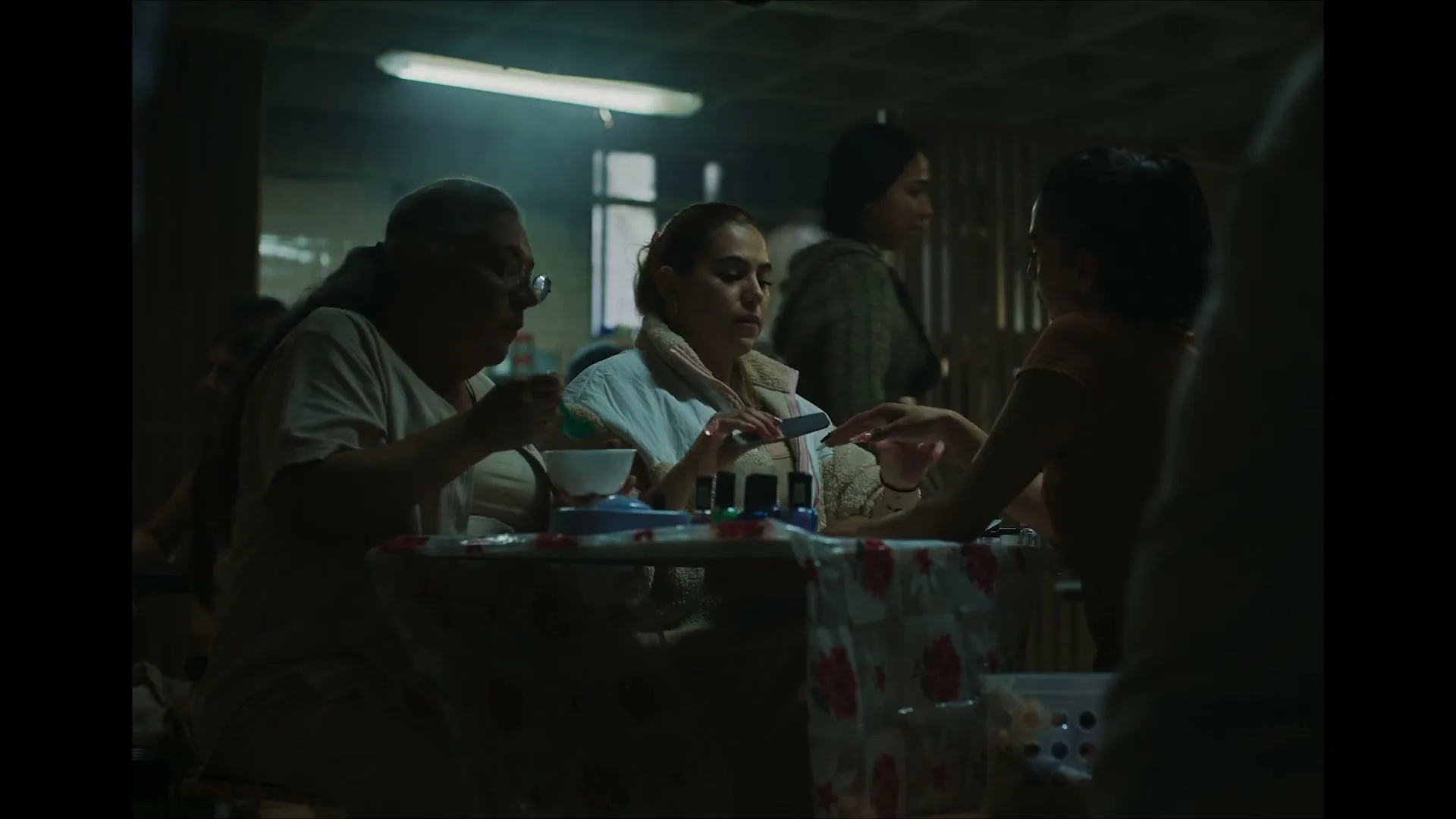 Three women are sitting at a table, one eating, one getting her nails done, and another watching, in a dimly lit room.
