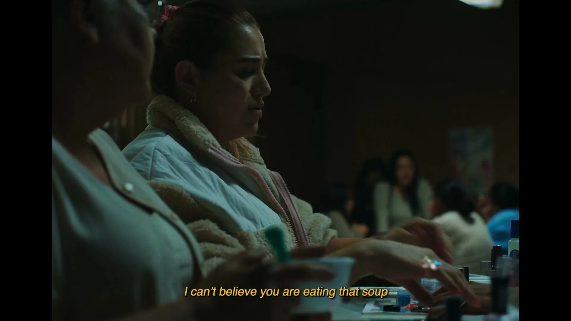 A woman with a pink scrunchie in her hair and a white jacket looks distressed while sitting at a table with other women, eating soup.