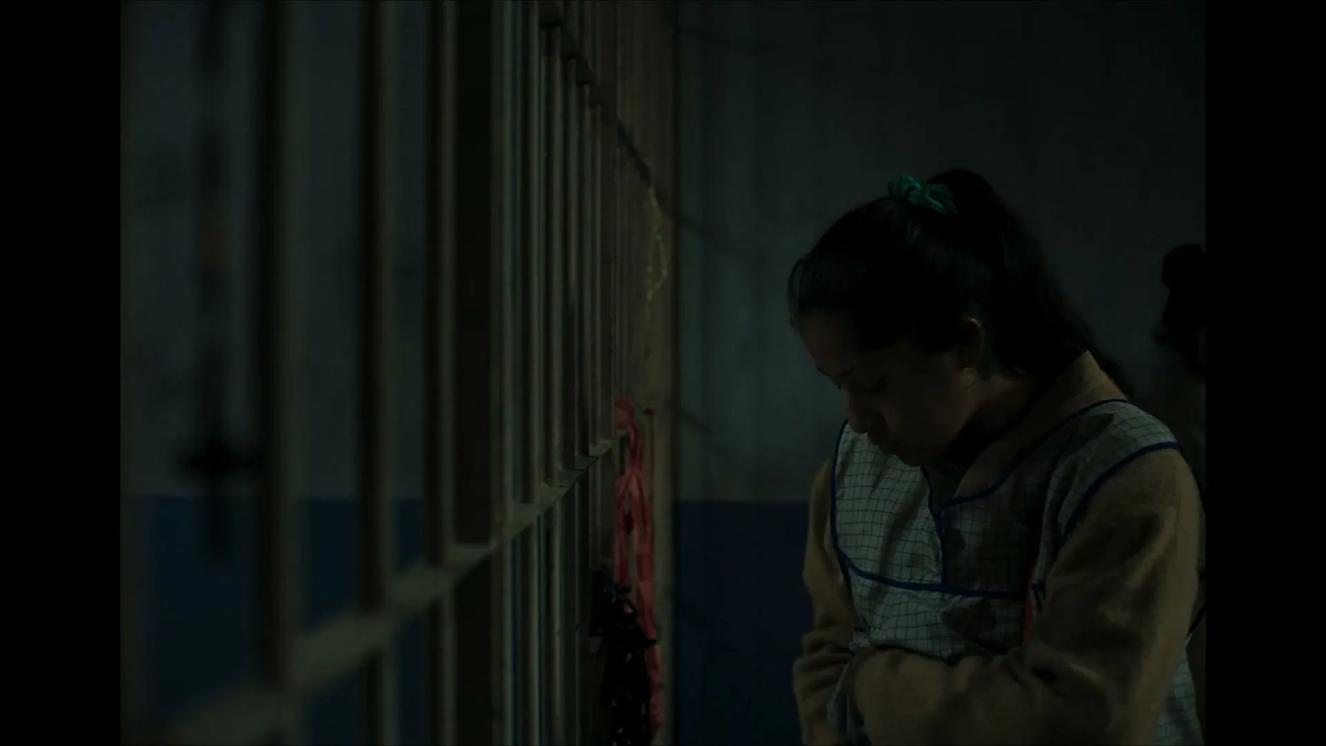 A young woman with dark hair in a ponytail, wearing a light blue and white checkered apron over a beige sweater, stands in a dimly lit prison cell, looking downcast.