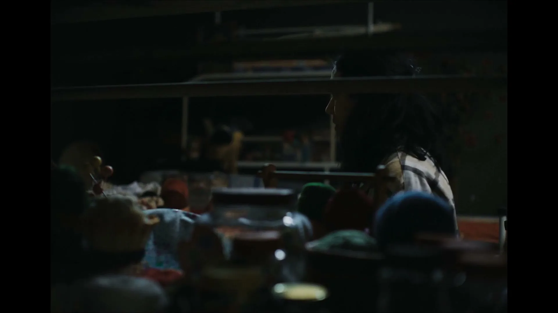 A young woman with dark hair and a patterned shirt looks at another woman in a dimly lit room, with various objects in the foreground.