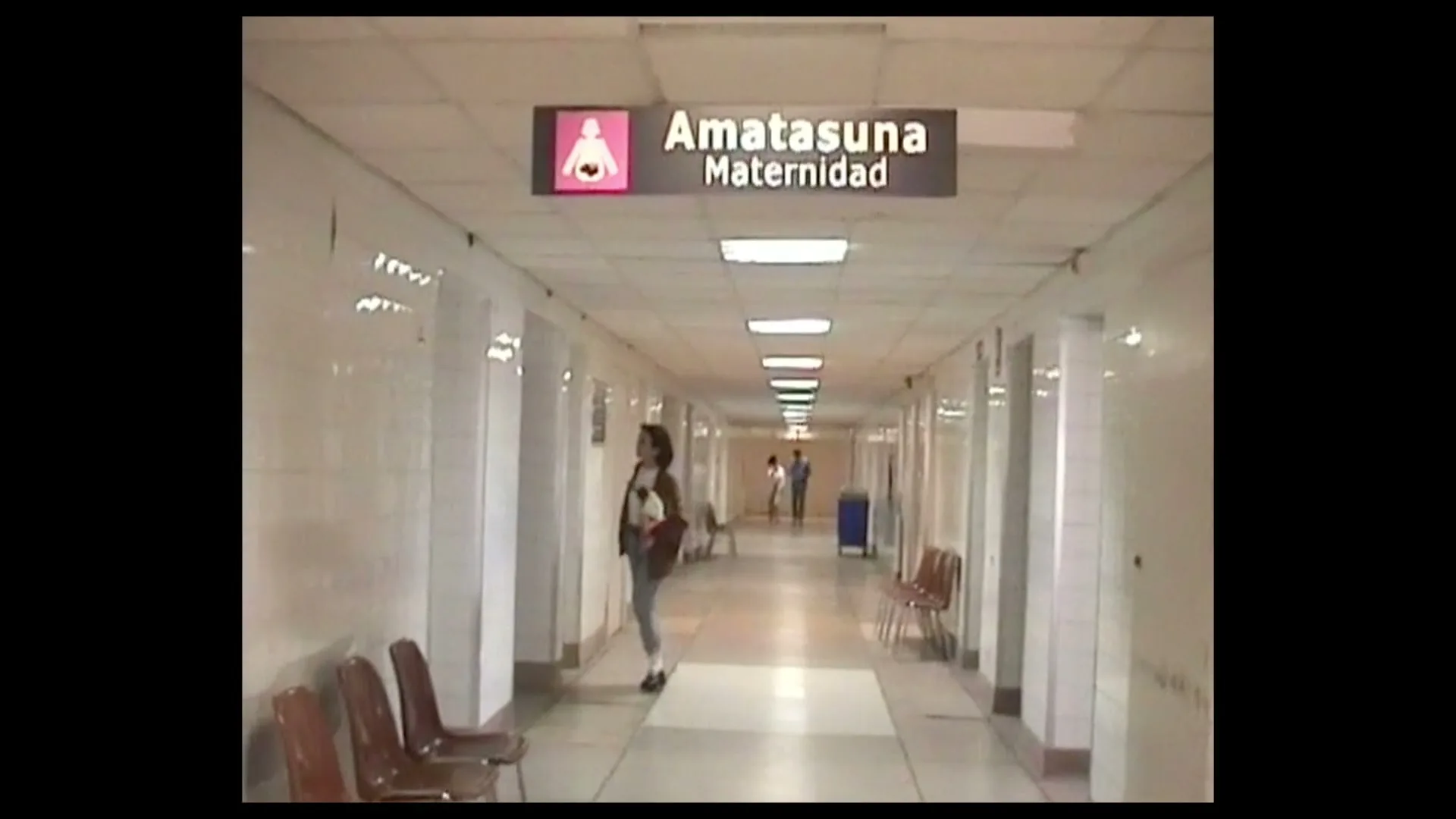 A woman in a white shirt and grey pants walks down a long, empty hospital corridor with a maternity sign overhead.