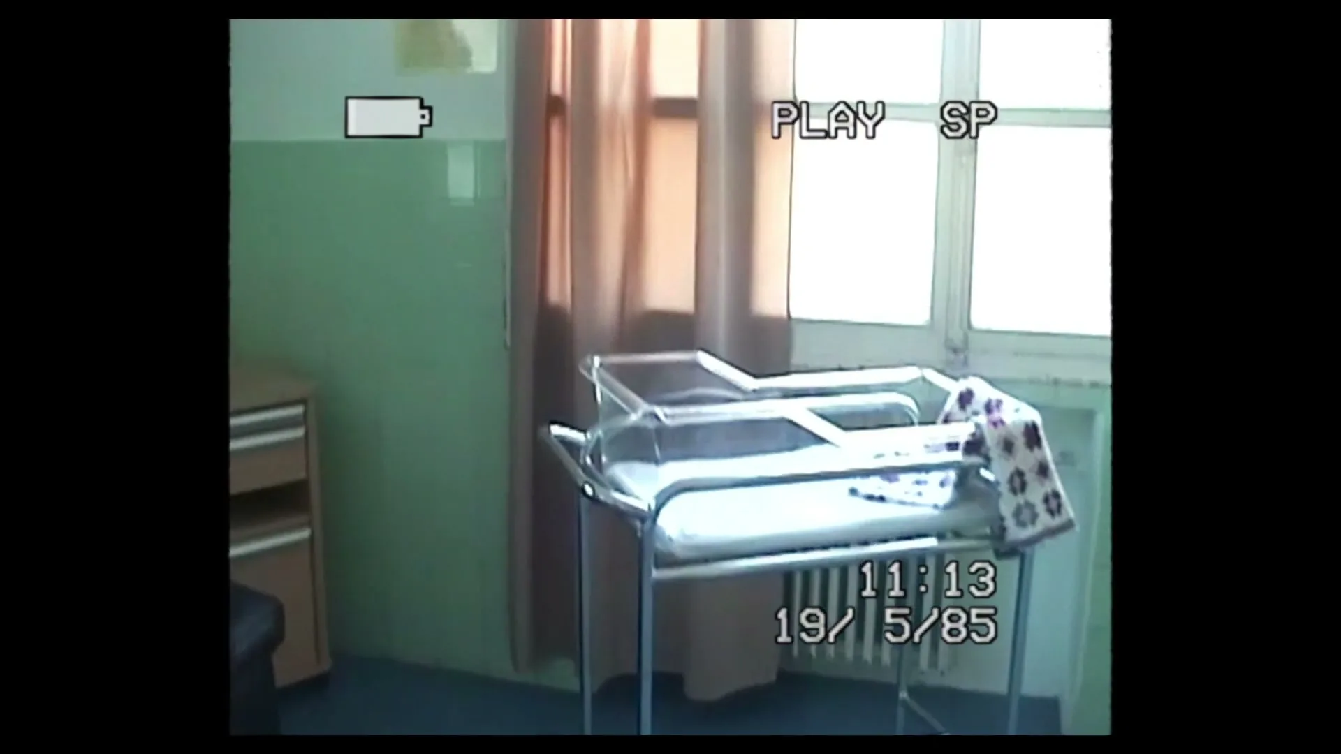 A vintage camcorder POV shot of an empty hospital room with a bassinet and a chair, with a window in the background.