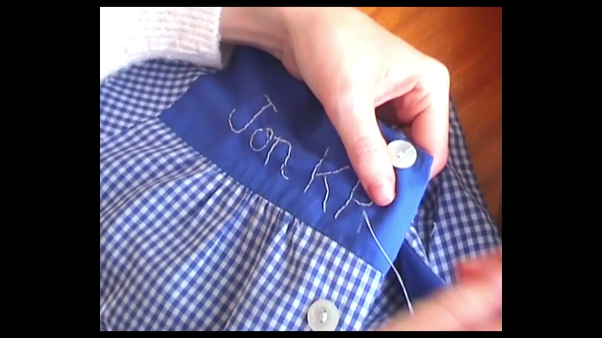 A close-up shot shows a person's hands sewing the name "Jon K" onto a blue and white checkered shirt, with a button visible.