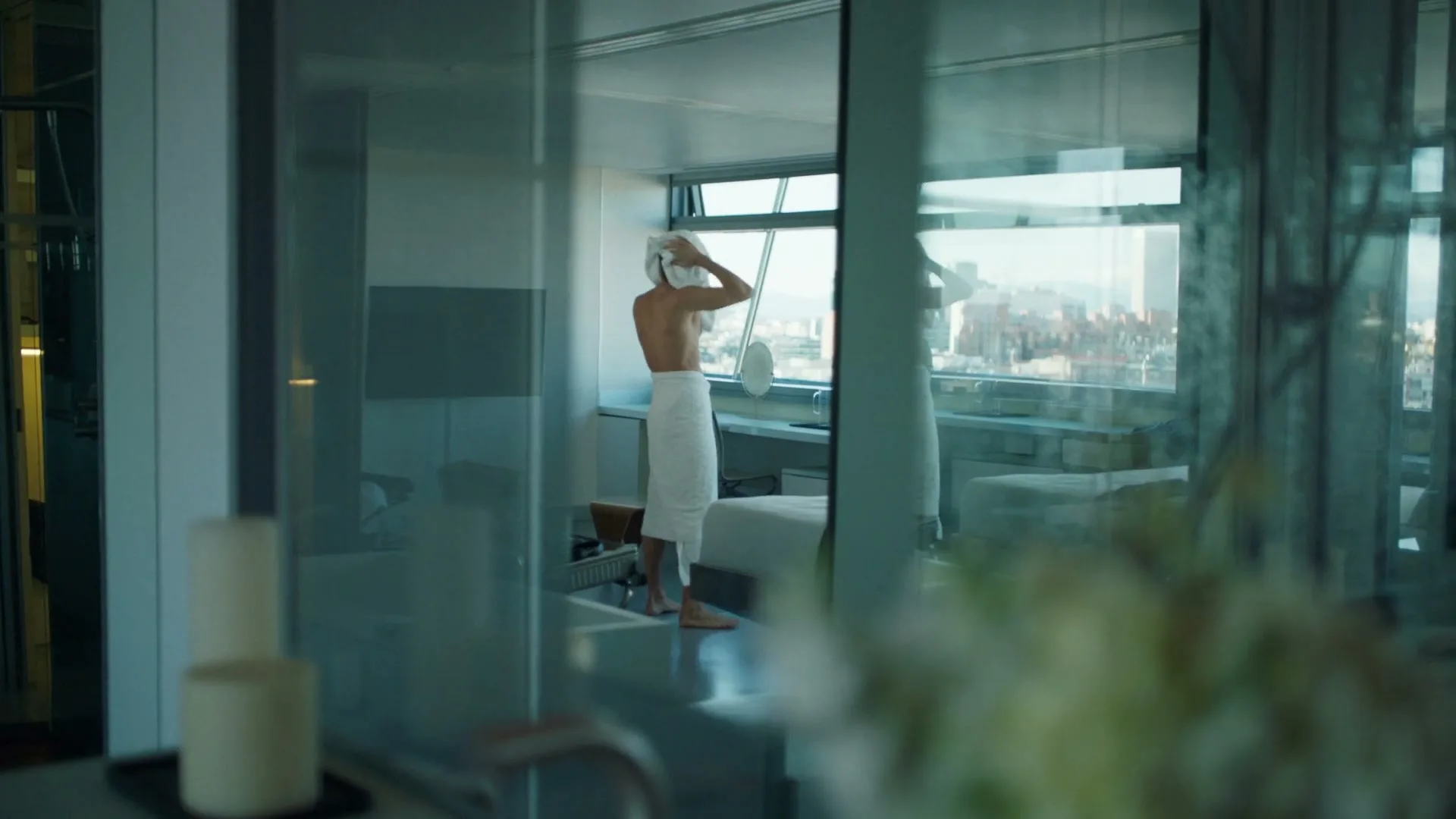 A man in his 30s, seen from behind, dries his hair with a white towel in a modern hotel room with a city view.