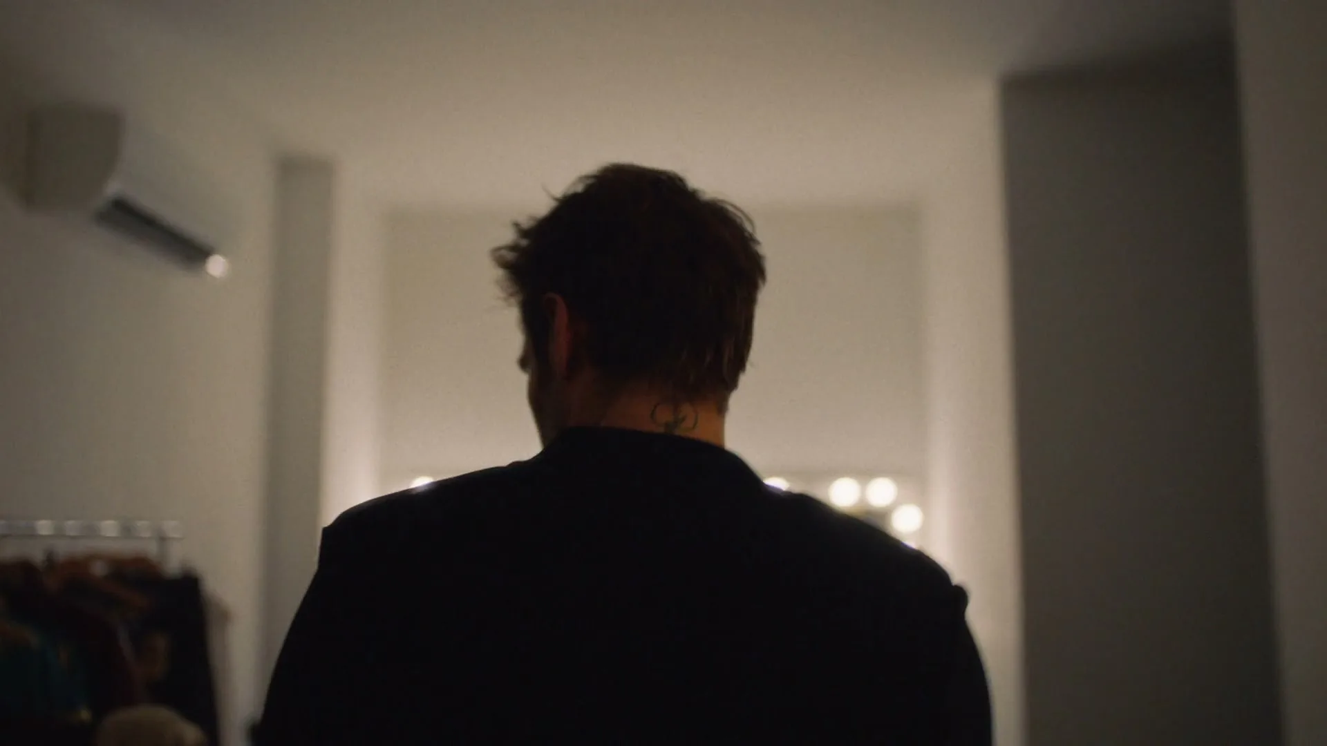 A man with dark, messy hair and a black jacket walks away from the camera in a dimly lit hallway towards a bright mirror.