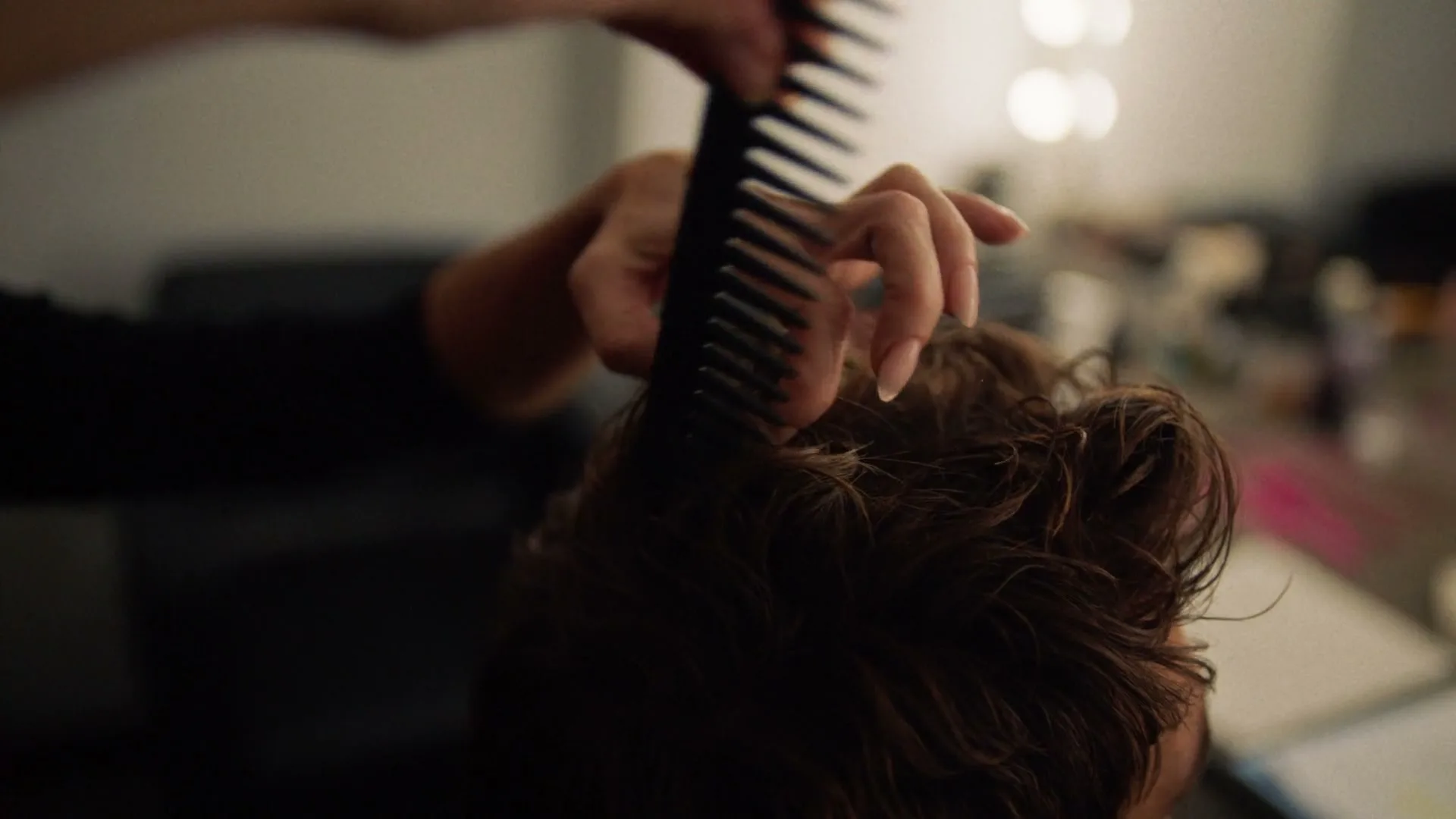 A person's hand combs through another person's wet, curly hair in a dimly lit interior setting.