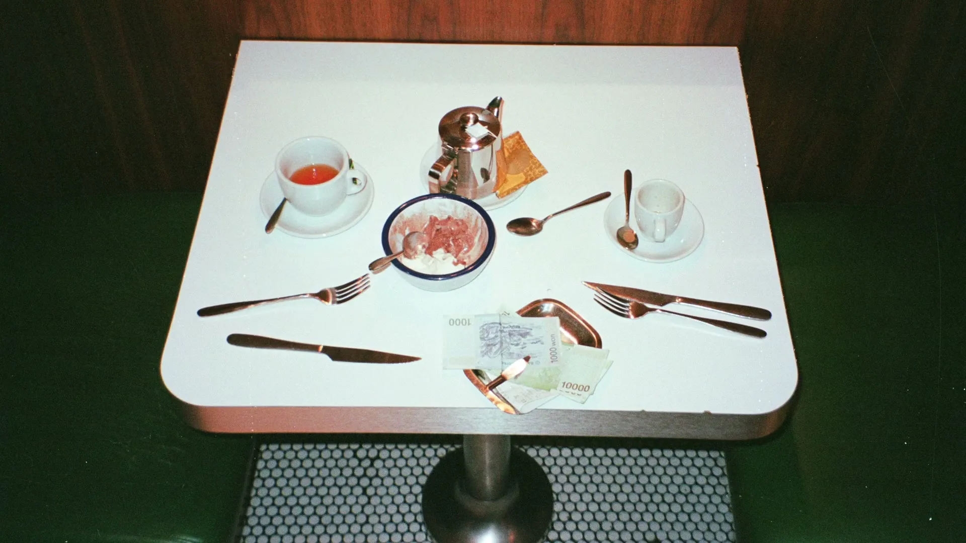 An overhead shot of a small white table set with cutlery, cups, a bowl of food, and money, in a restaurant booth.