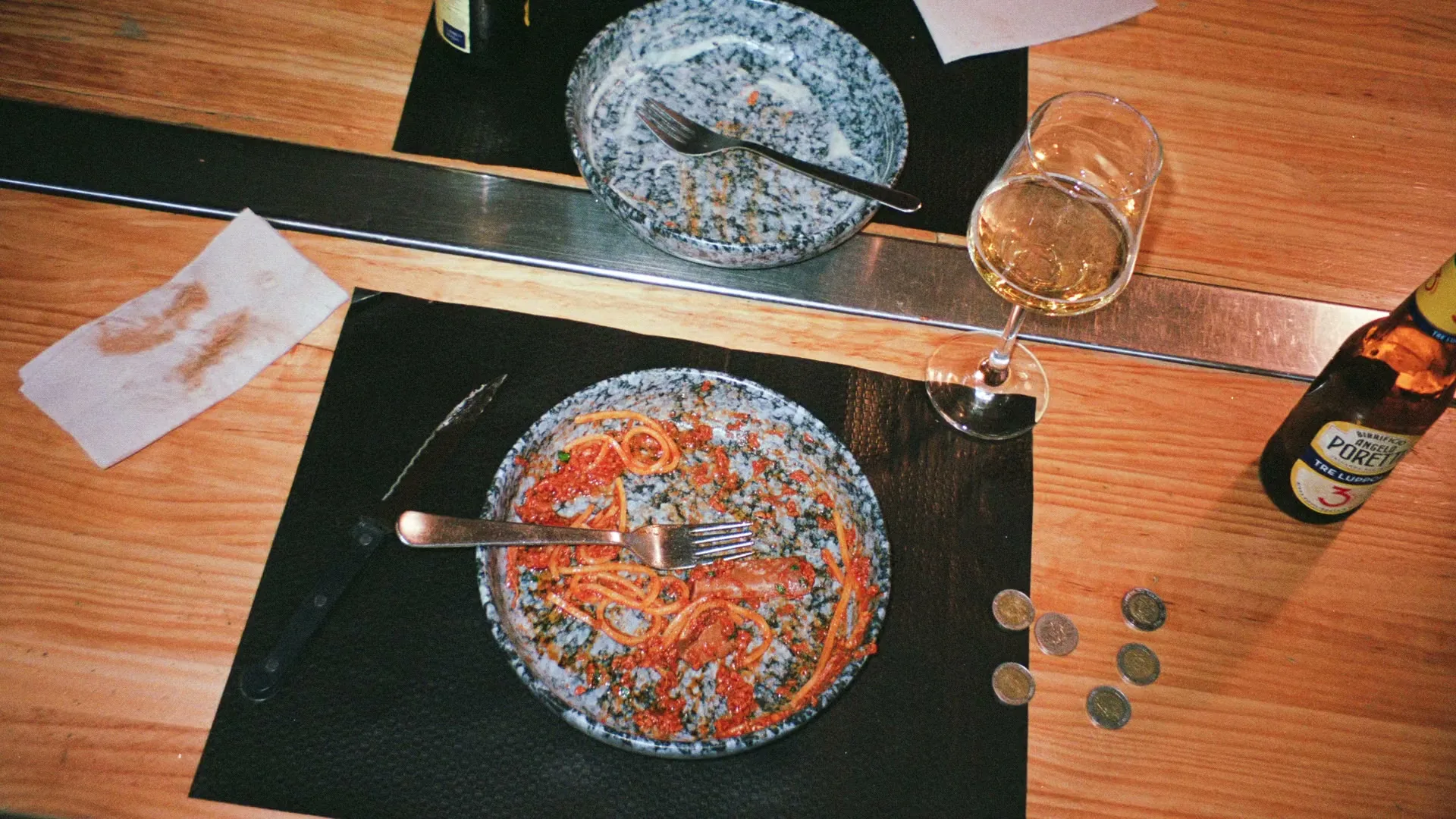An overhead shot of a wooden table with two plates of half-eaten pasta, a glass of white wine, a beer bottle, and some coins.