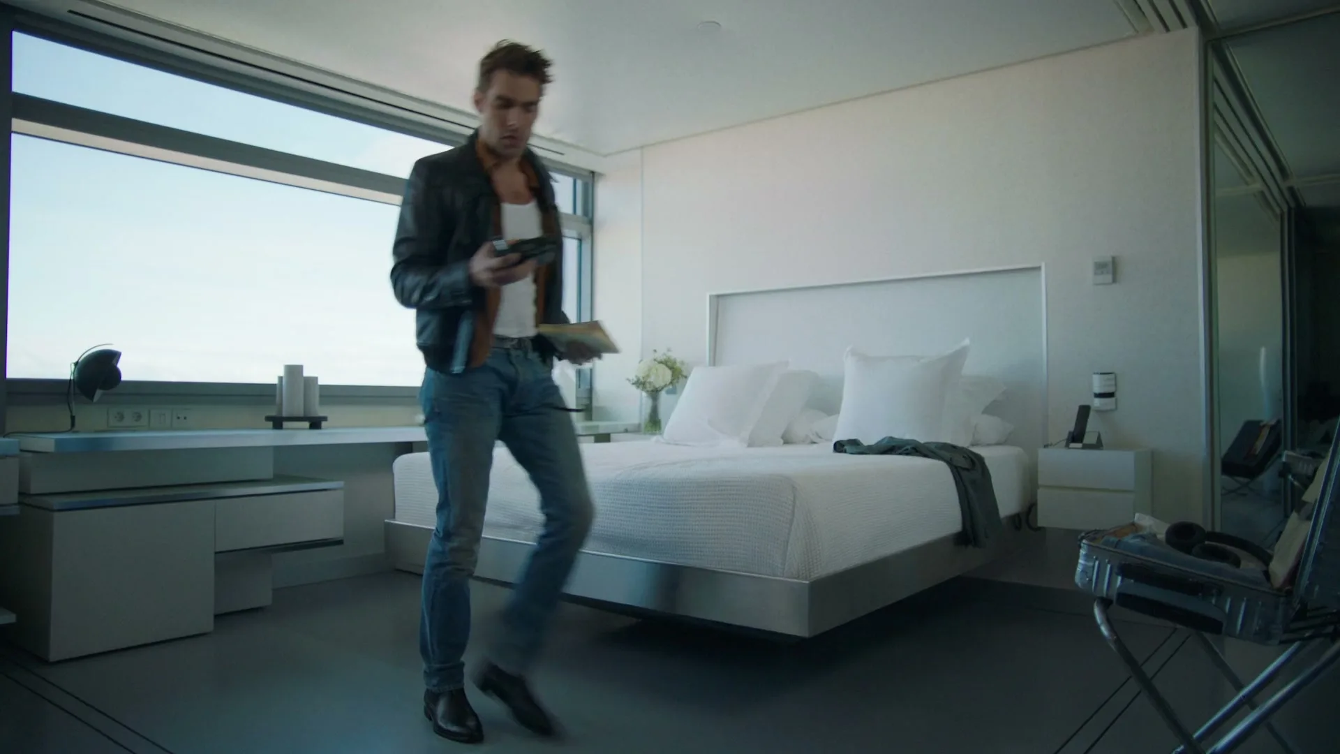 A young man in a leather jacket and jeans walks across a modern, minimalist bedroom, holding a phone and a book.