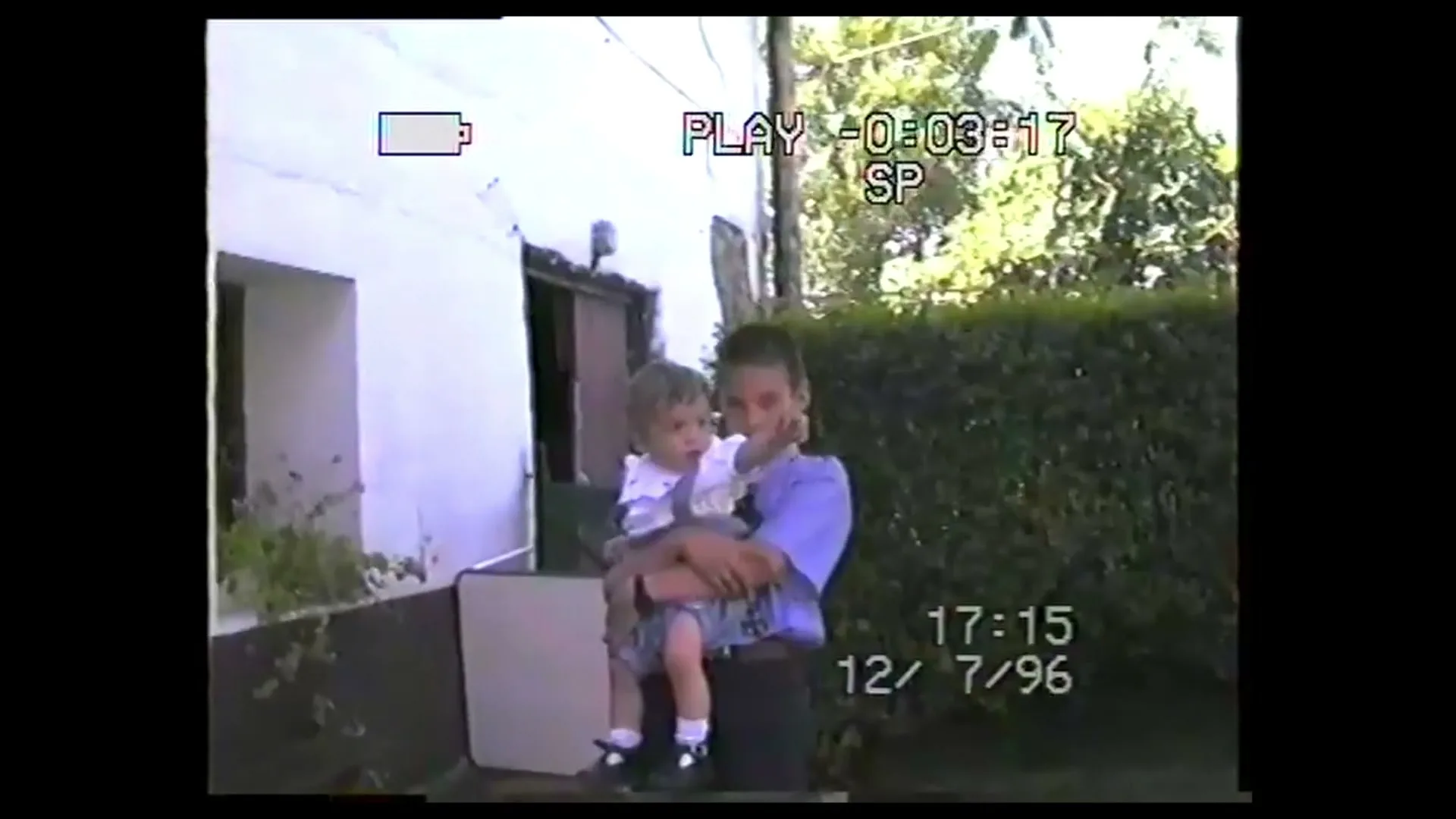 A young boy in a blue shirt holds a baby in a white shirt, both looking at the camera in a backyard setting.