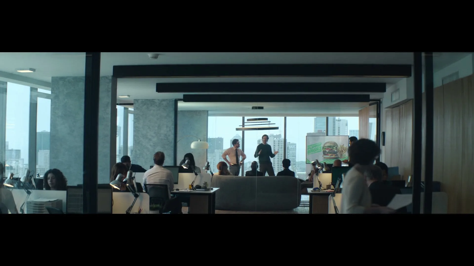A diverse group of office workers are seen in a modern office space with large windows overlooking a city, as two men present in front of a screen.