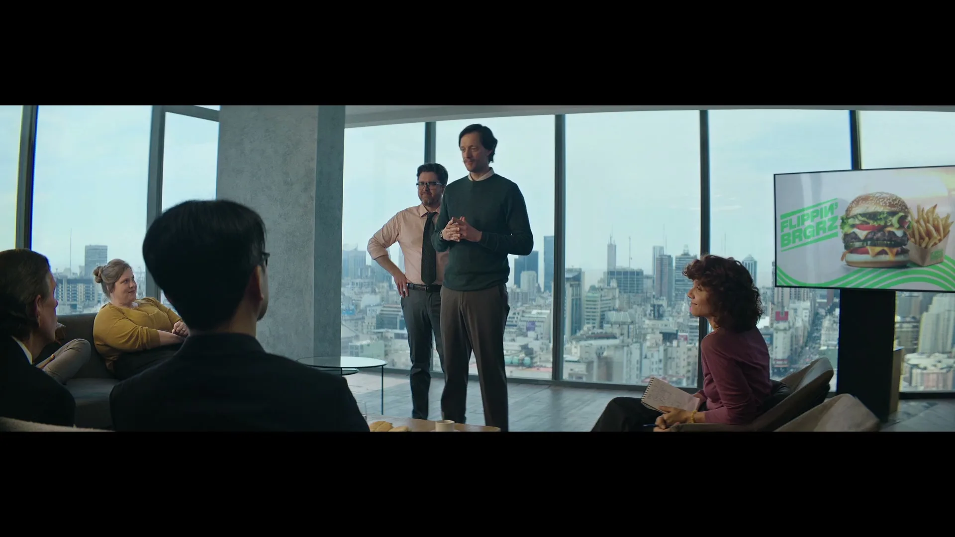 Two men present to a group of colleagues in a modern office with a city view, showcasing a burger and fries on a large screen.