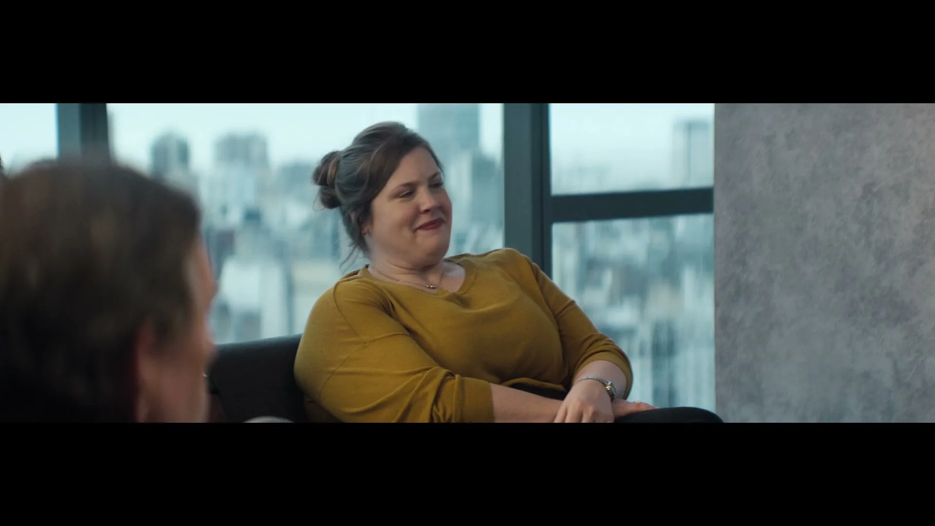A woman in her 30s, wearing a mustard yellow sweater, sits in an office chair, smiling and looking to her left, with a city skyline visible through a large window behind her.