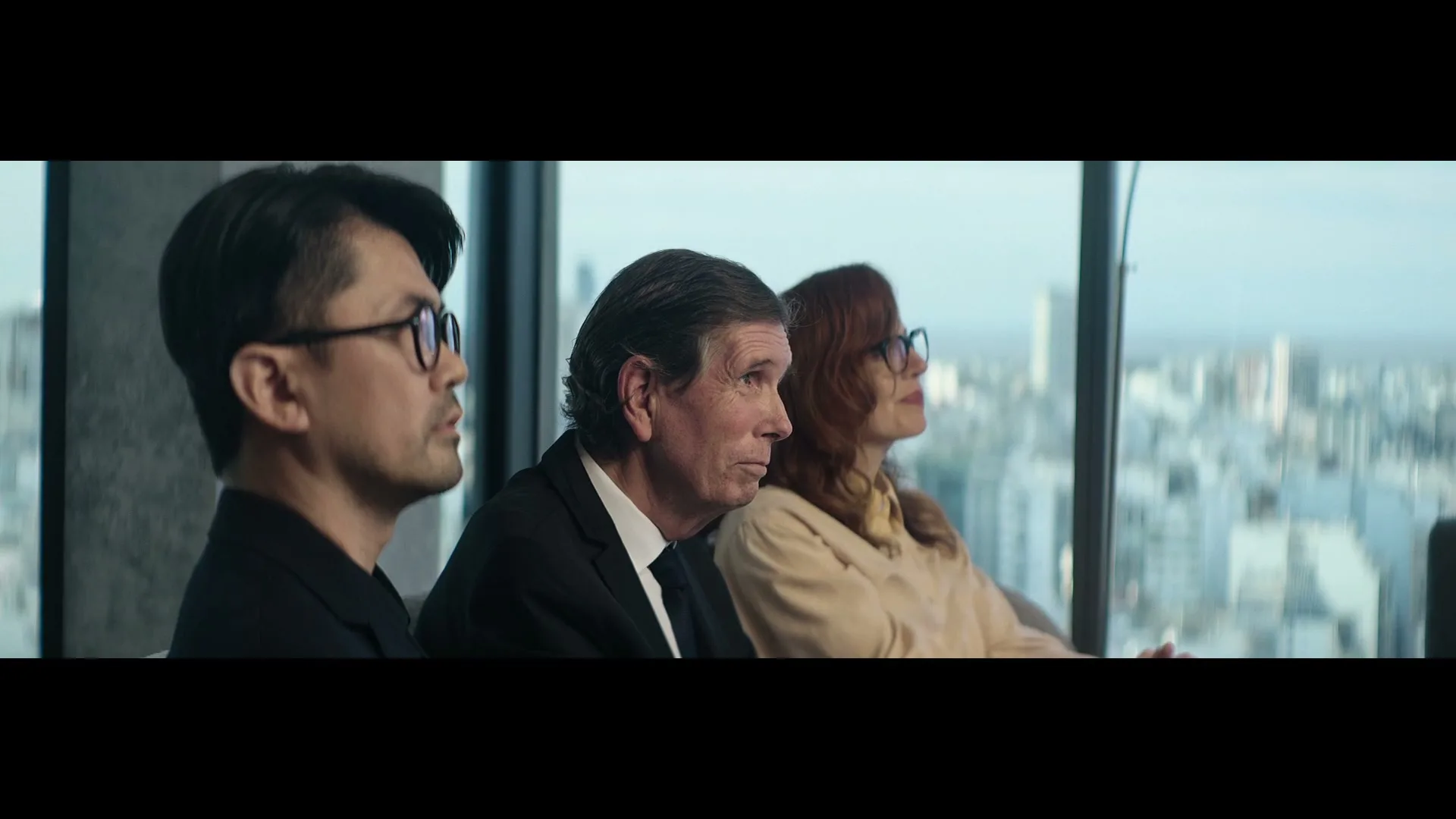 Three business people, two men and one woman, sit in a row looking out a window at a city skyline.