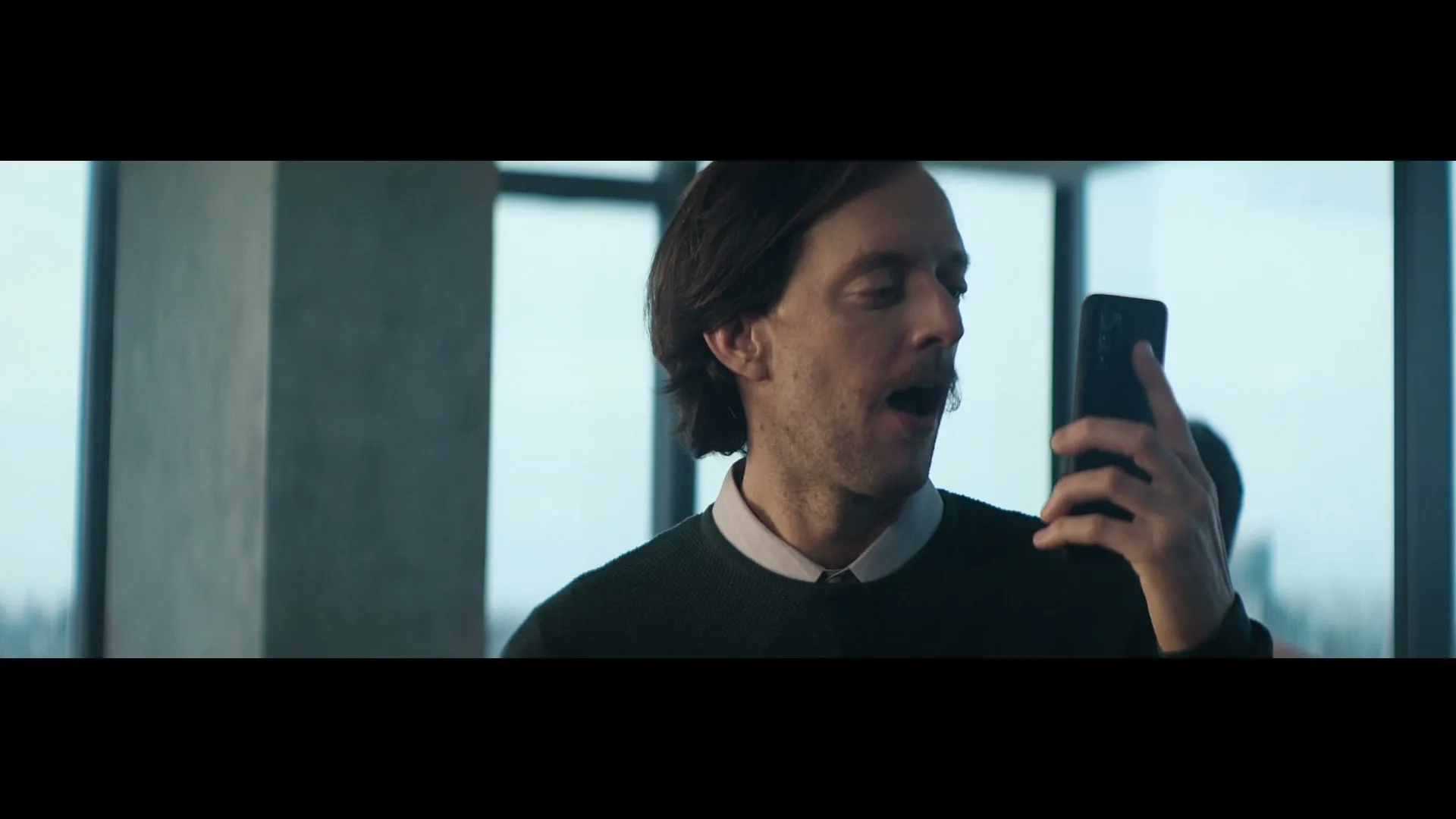 A man with a mustache holds a smartphone to his mouth, then looks surprised, while another man is blurred in the background in an office.