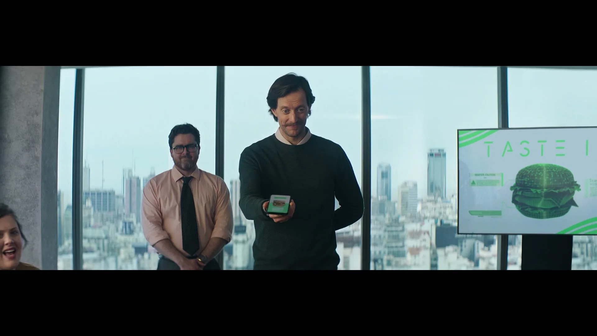 A man in a dark sweater holds a small device, presenting to a group in a modern office with a city skyline view, while a woman reaches for the device.