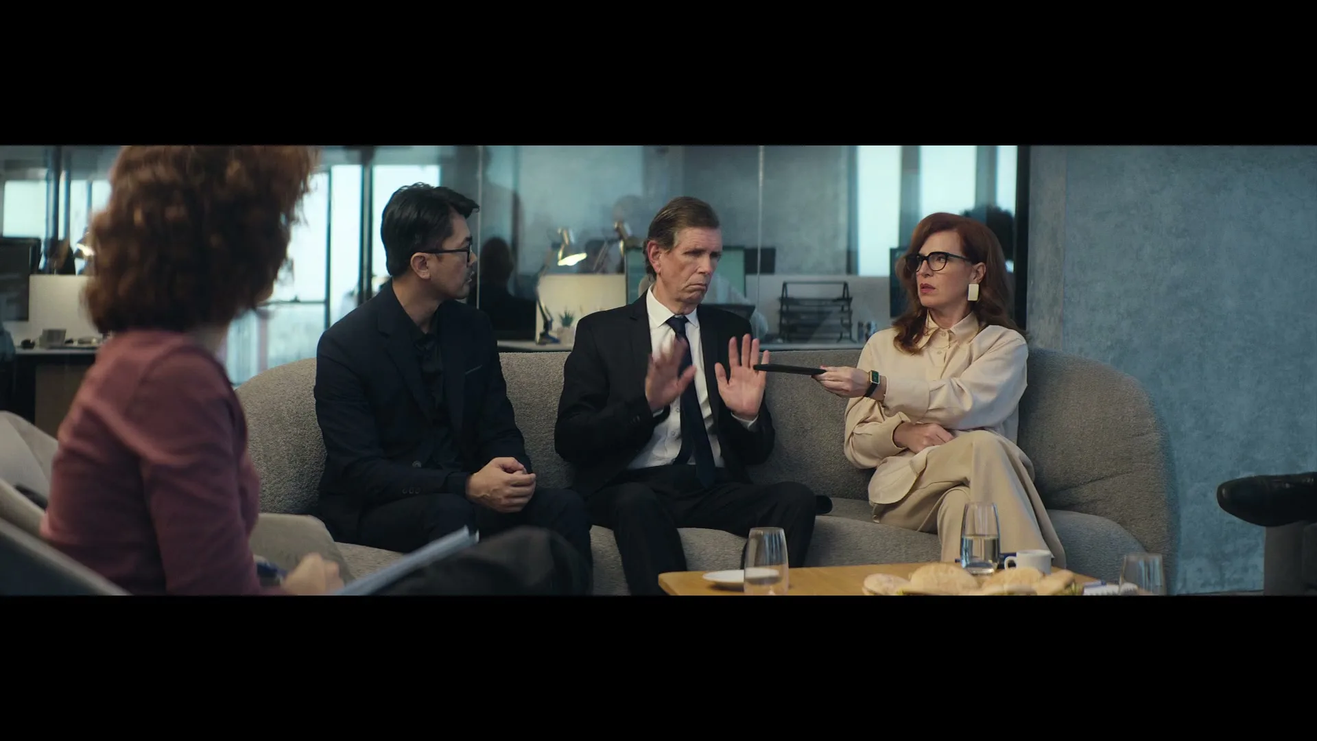 Four business people sit on a couch in an office, with a man in the middle gesturing with his hands.