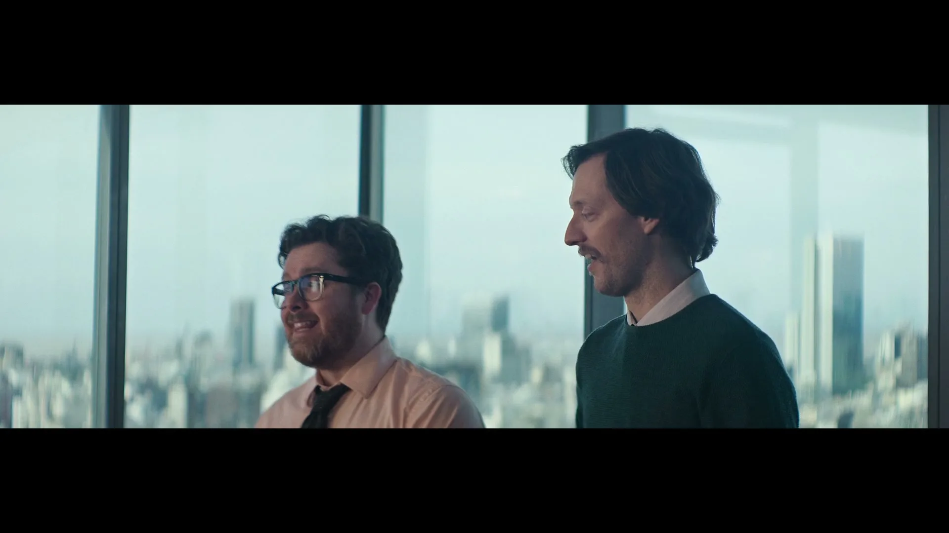 Two men, one with glasses and a beard, the other with a mustache, stand in an office with a city view, reacting with amusement.