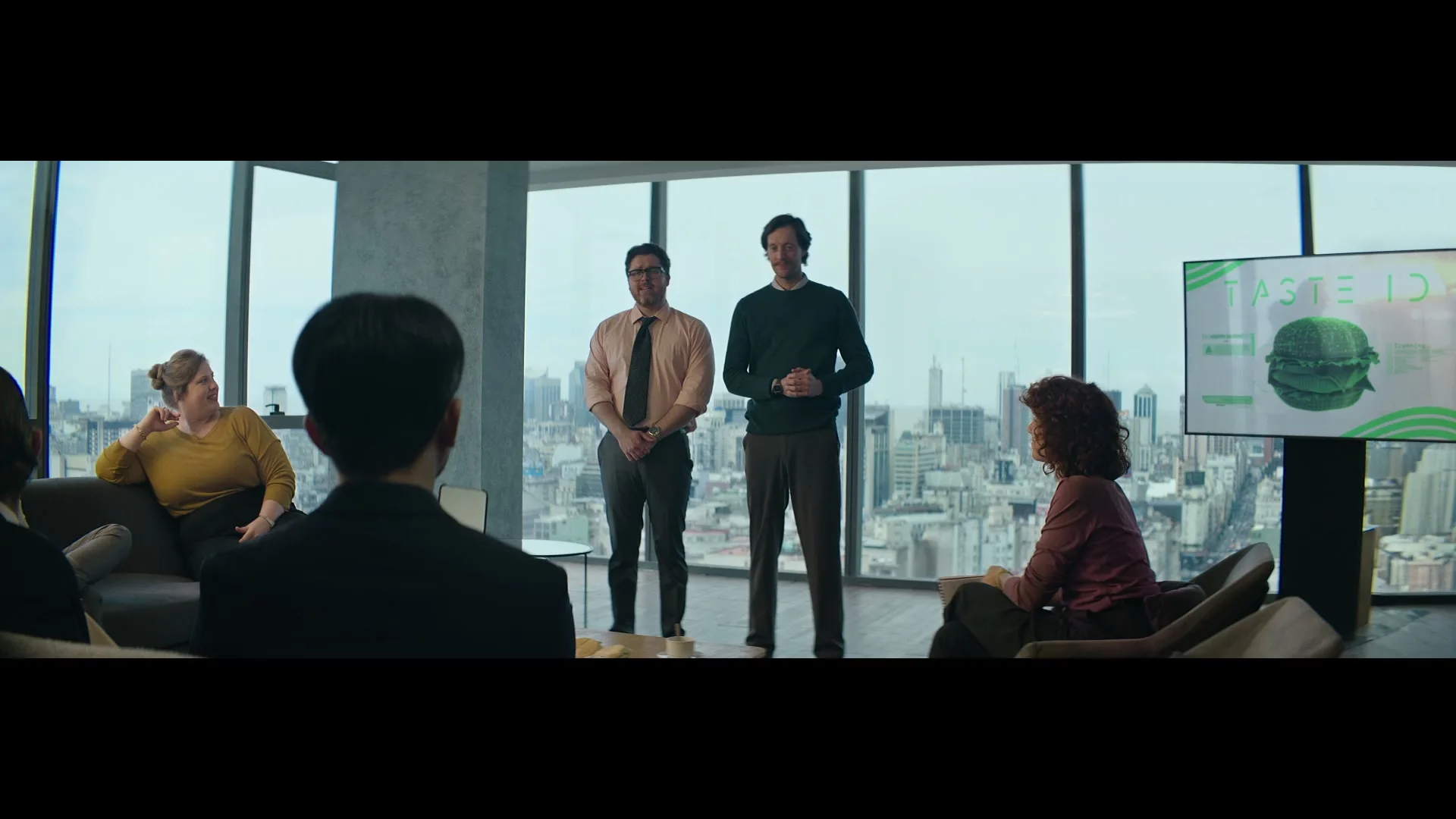 Two men present a "Taste ID" project with a green burger on screen to a group of colleagues in a modern high-rise office with a city view.