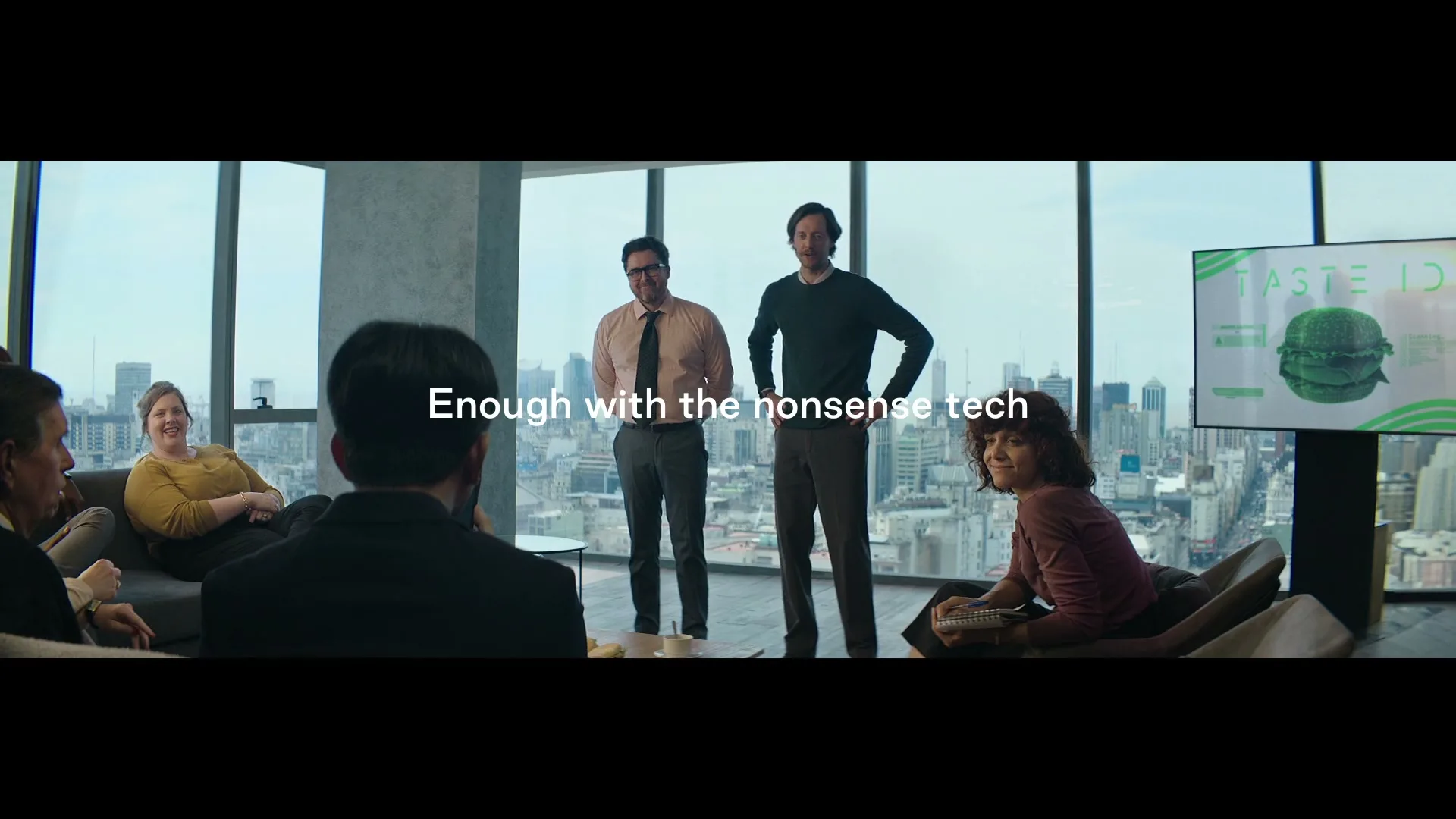 Two men present a "Taste ID" burger on a screen to a group of people in a modern office with a city view.