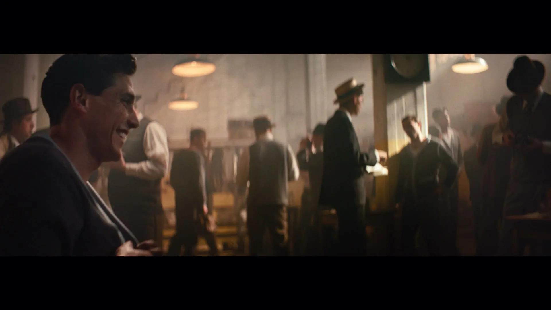 A man in a dark shirt smiles while looking off-camera, surrounded by other men in a dimly lit, smoky room.