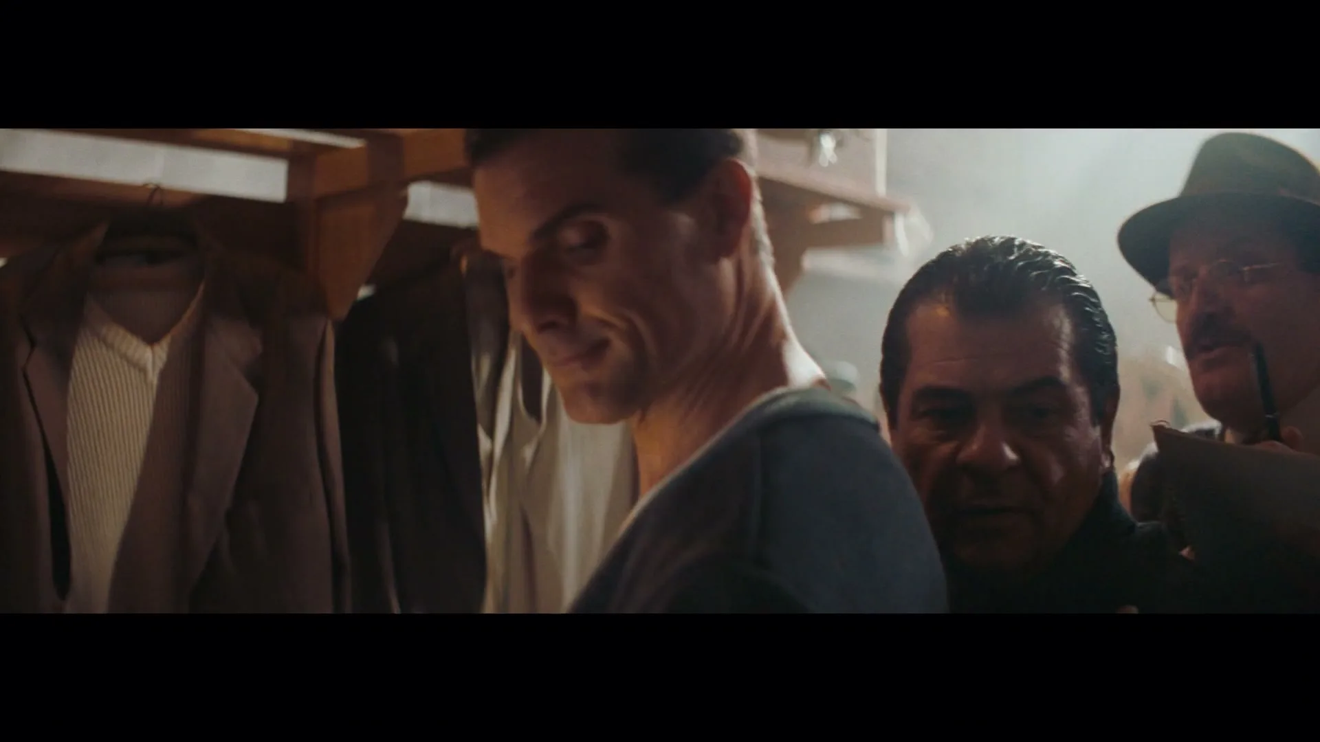 A male athlete in a locker room turns to smile at the camera, while other men are visible in the background.