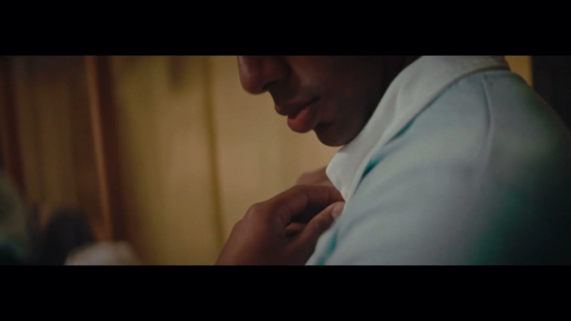 A man in his 20s, wearing a light blue shirt, adjusts his collar in a dimly lit room, preparing for an important event.