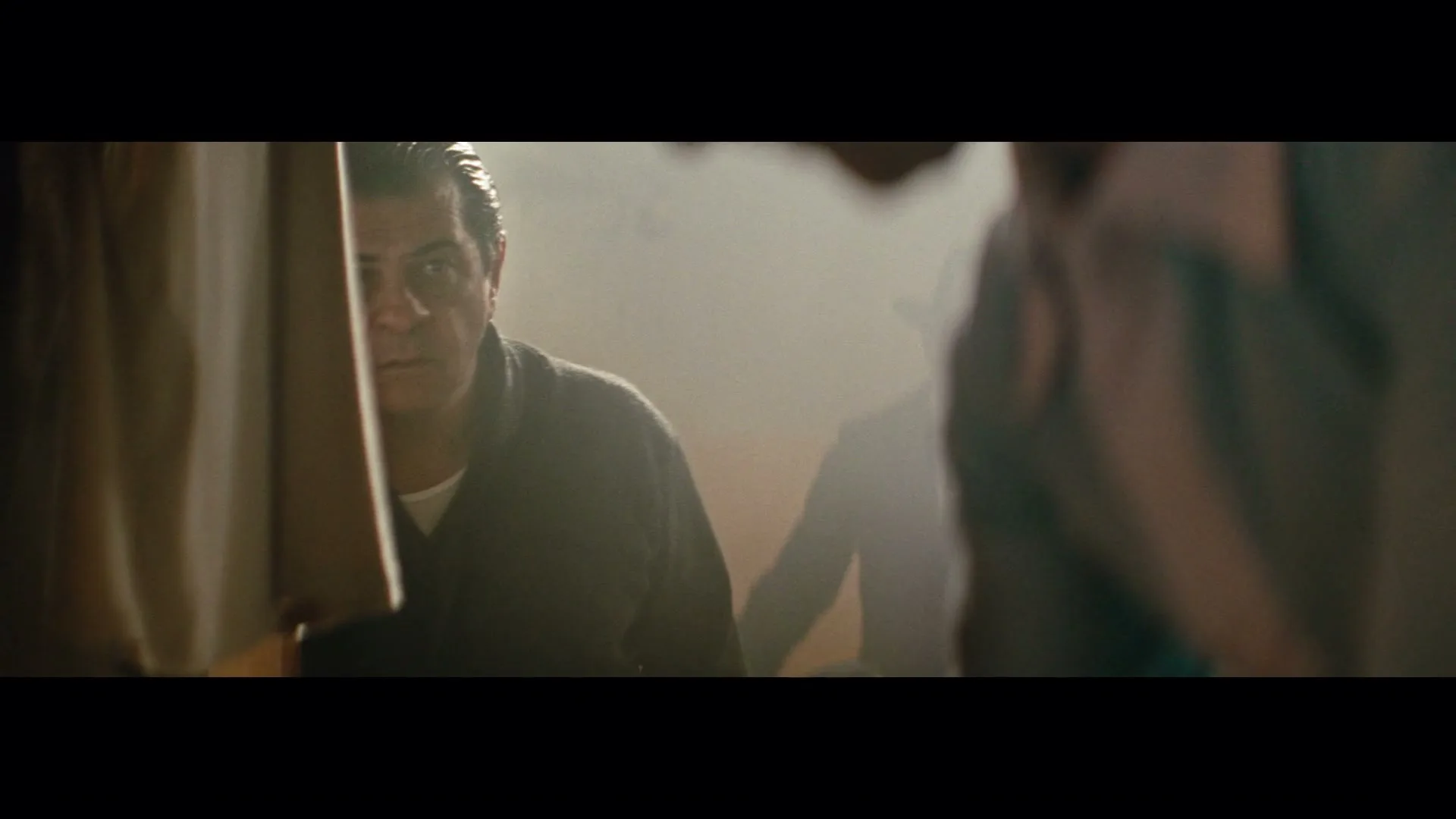 A middle-aged man with dark hair and a dark robe looks intently from behind a curtain, with other figures blurred in the smoky background.