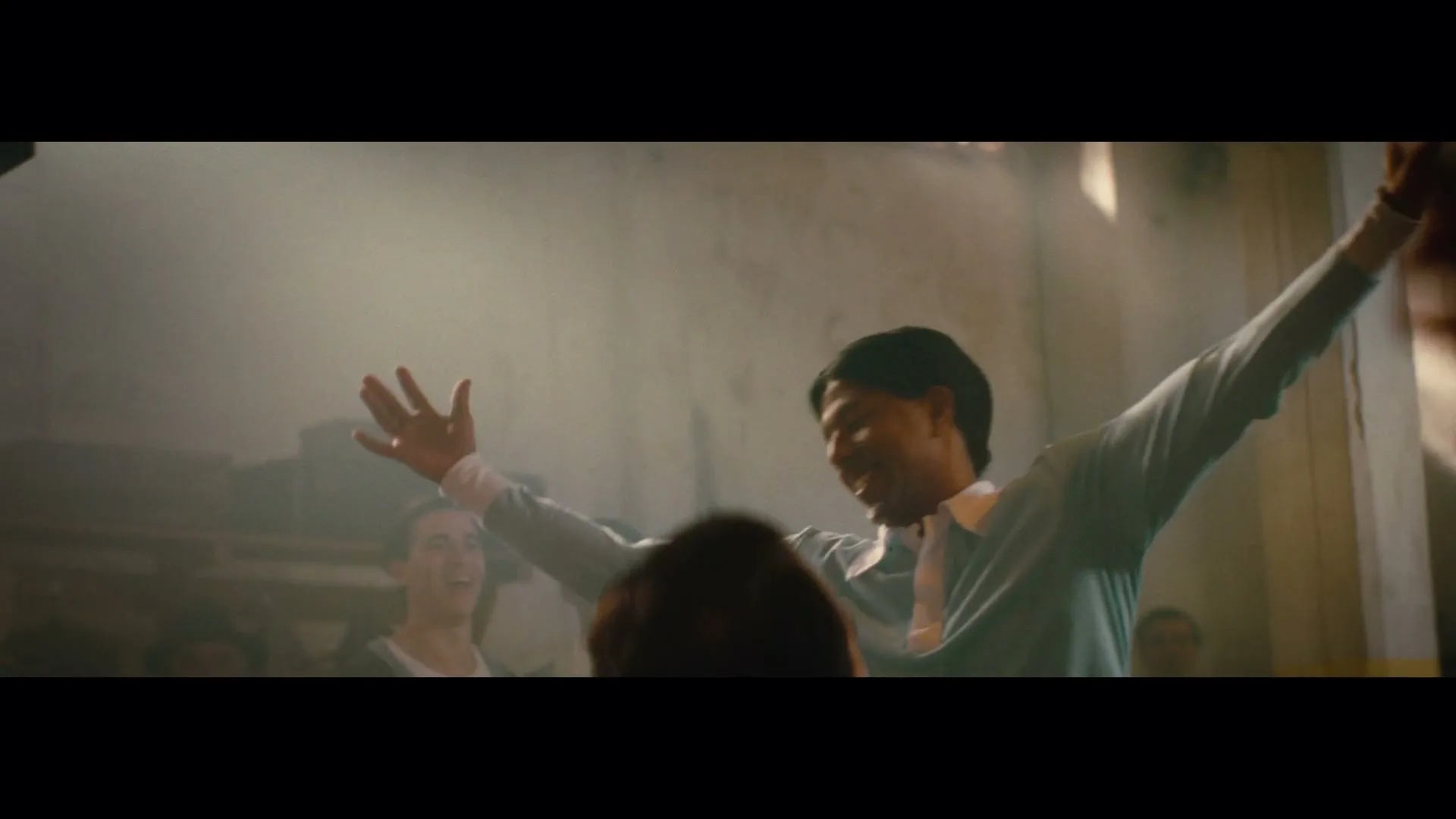 A man in a light blue shirt and tie raises his arms in triumph, smiling joyfully amidst a cheering crowd in a smoky, dimly lit room.