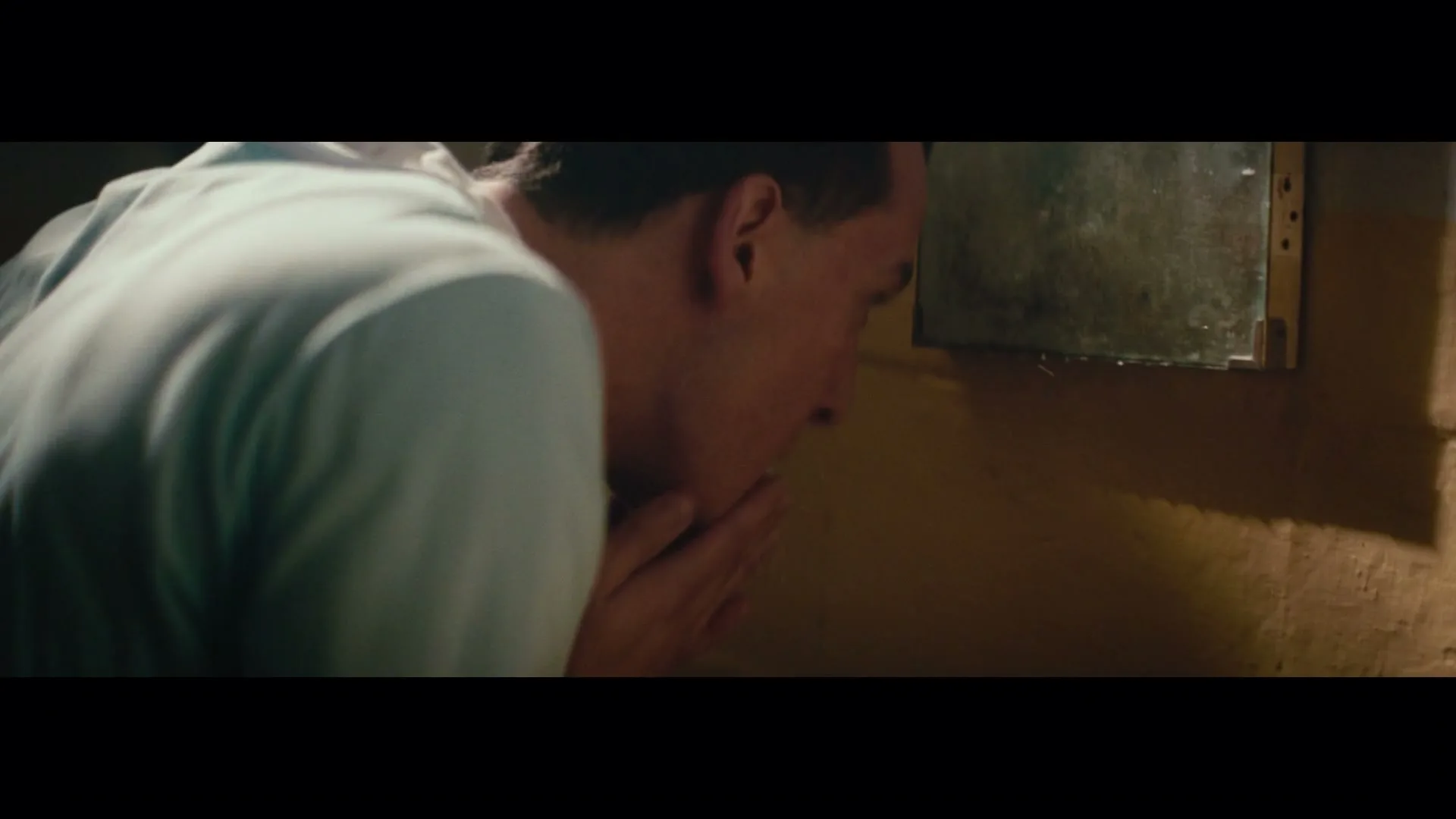 A male athlete in a light blue shirt splashes water on his face from a small, rustic sink in a dimly lit locker room.