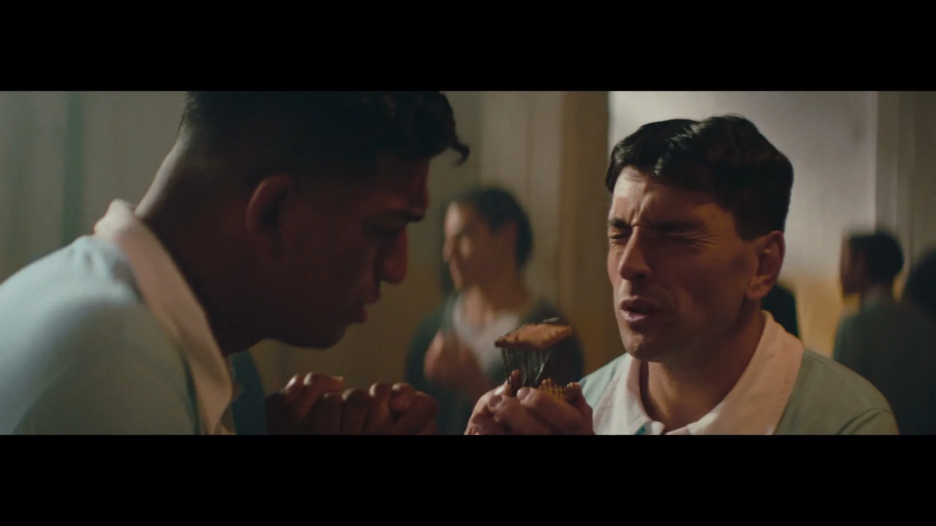 Two male soccer players in light blue jerseys share a mate gourd in a locker room, one with a pained expression.