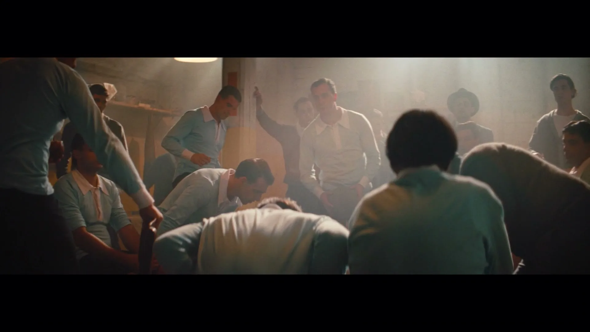 A group of male soccer players in vintage uniforms huddle together in a dimly lit locker room, some bending over, others looking around.