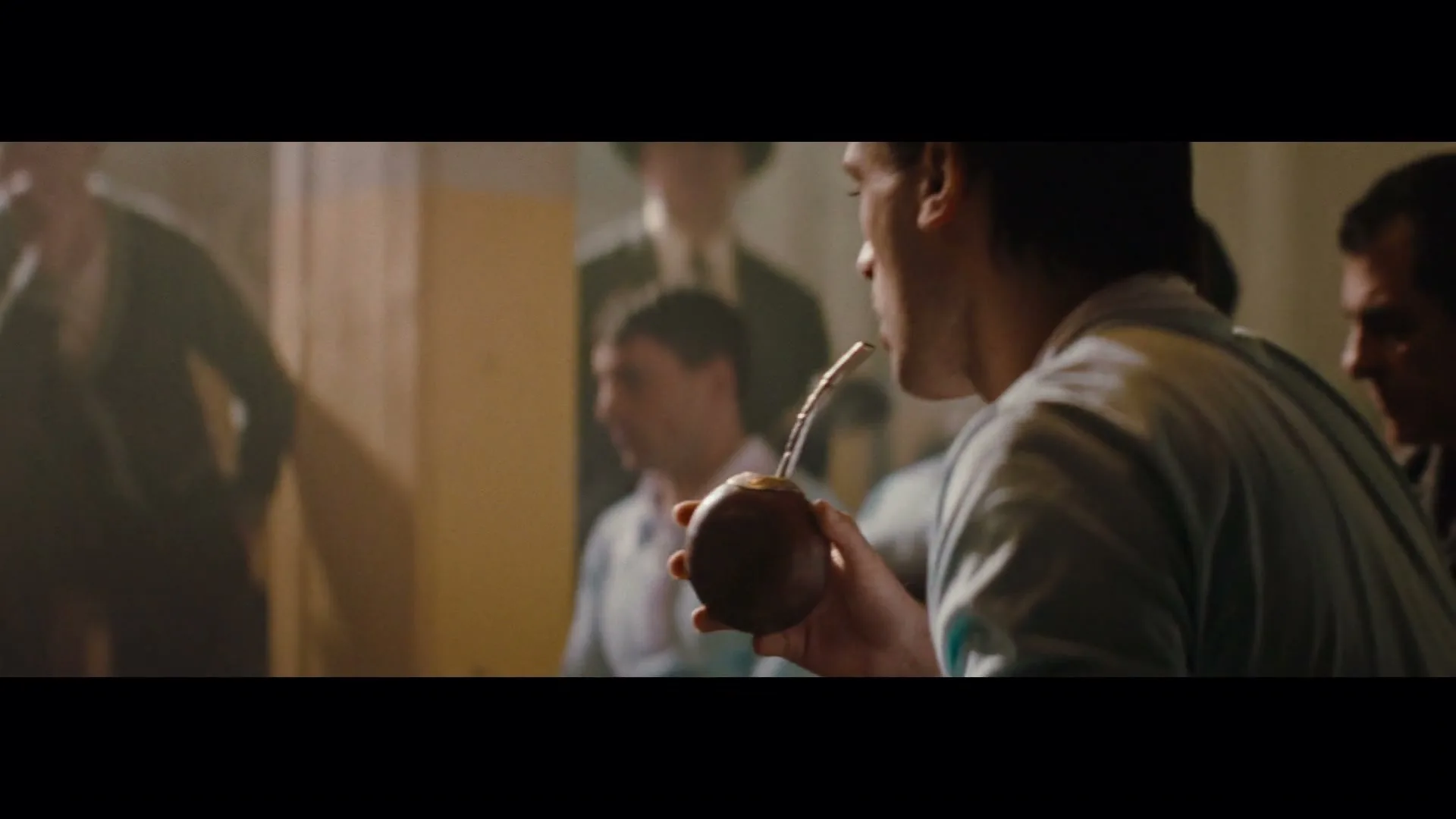 A man in a light blue shirt passes a mate gourd to another man, who then drinks from it, surrounded by other men in a dimly lit room.