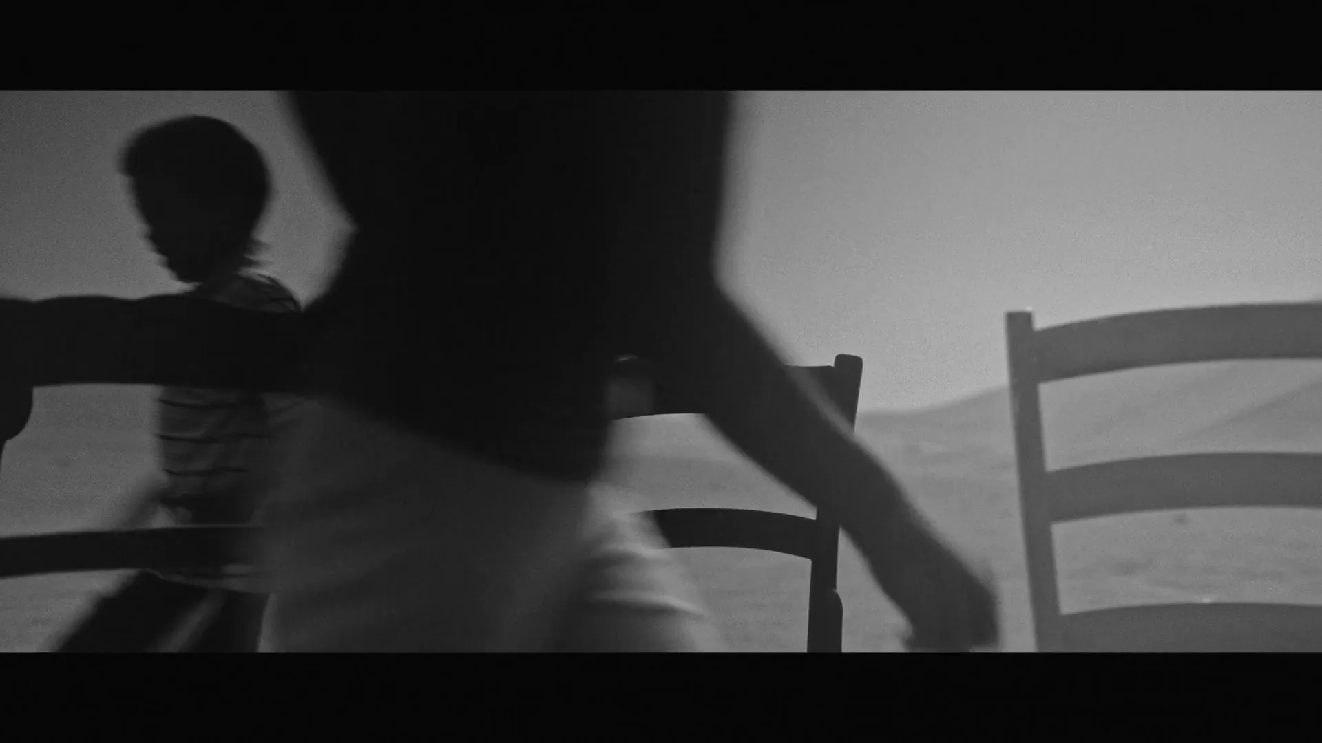 Three children run past empty chairs in a desolate, dusty landscape, their figures silhouetted against the bright sky in black and white.