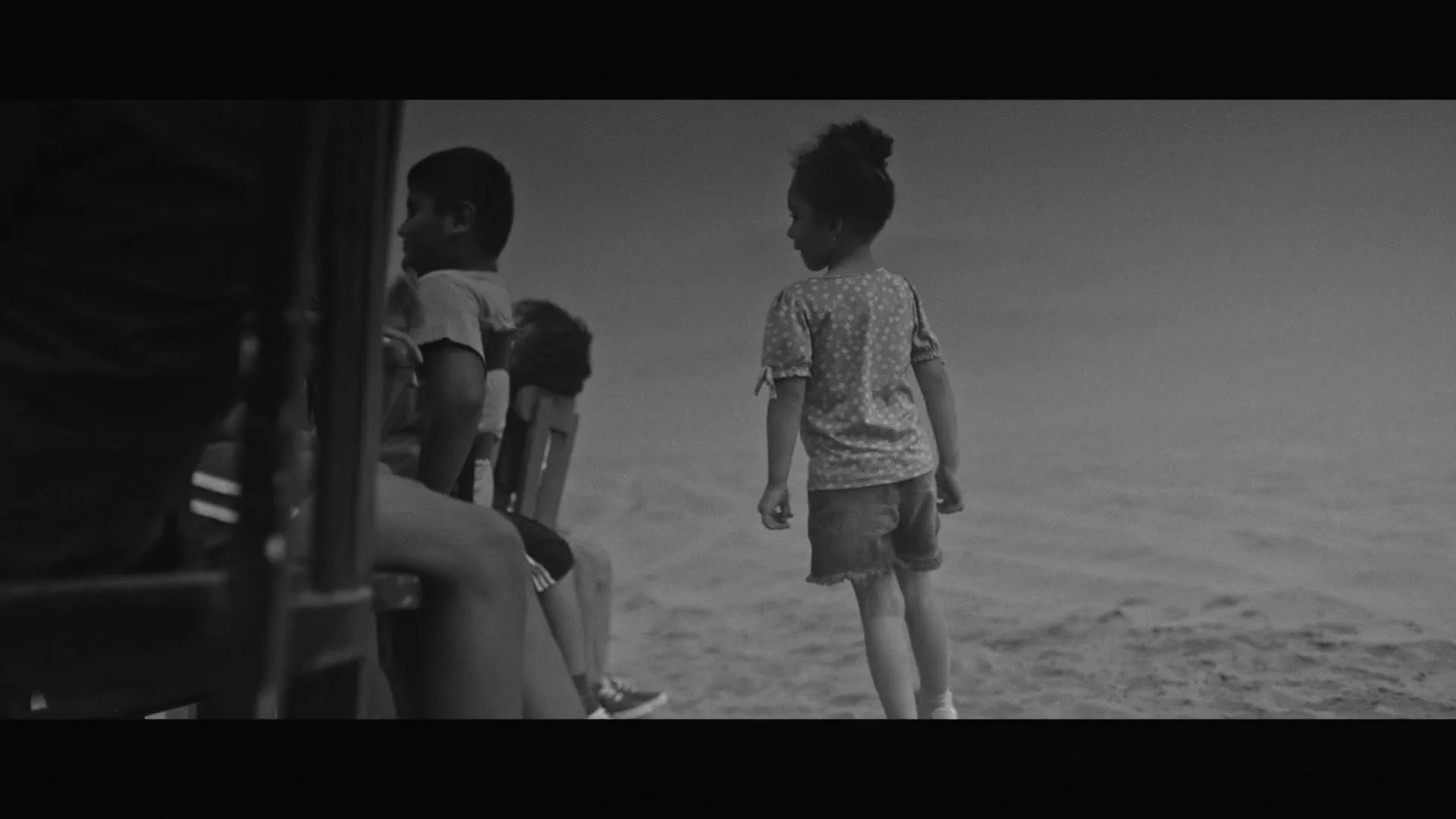 Two children, a boy and a girl, stand on a sandy beach, looking out at the water, in a black and white shot.
