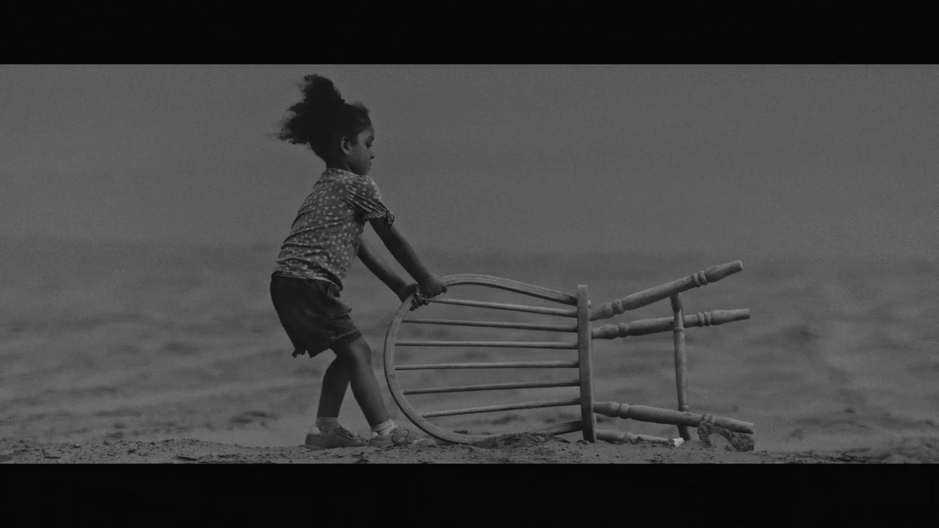 A young girl in a floral shirt and shorts pushes a wooden chair across a sandy, barren landscape, then other children run to sit on chairs.