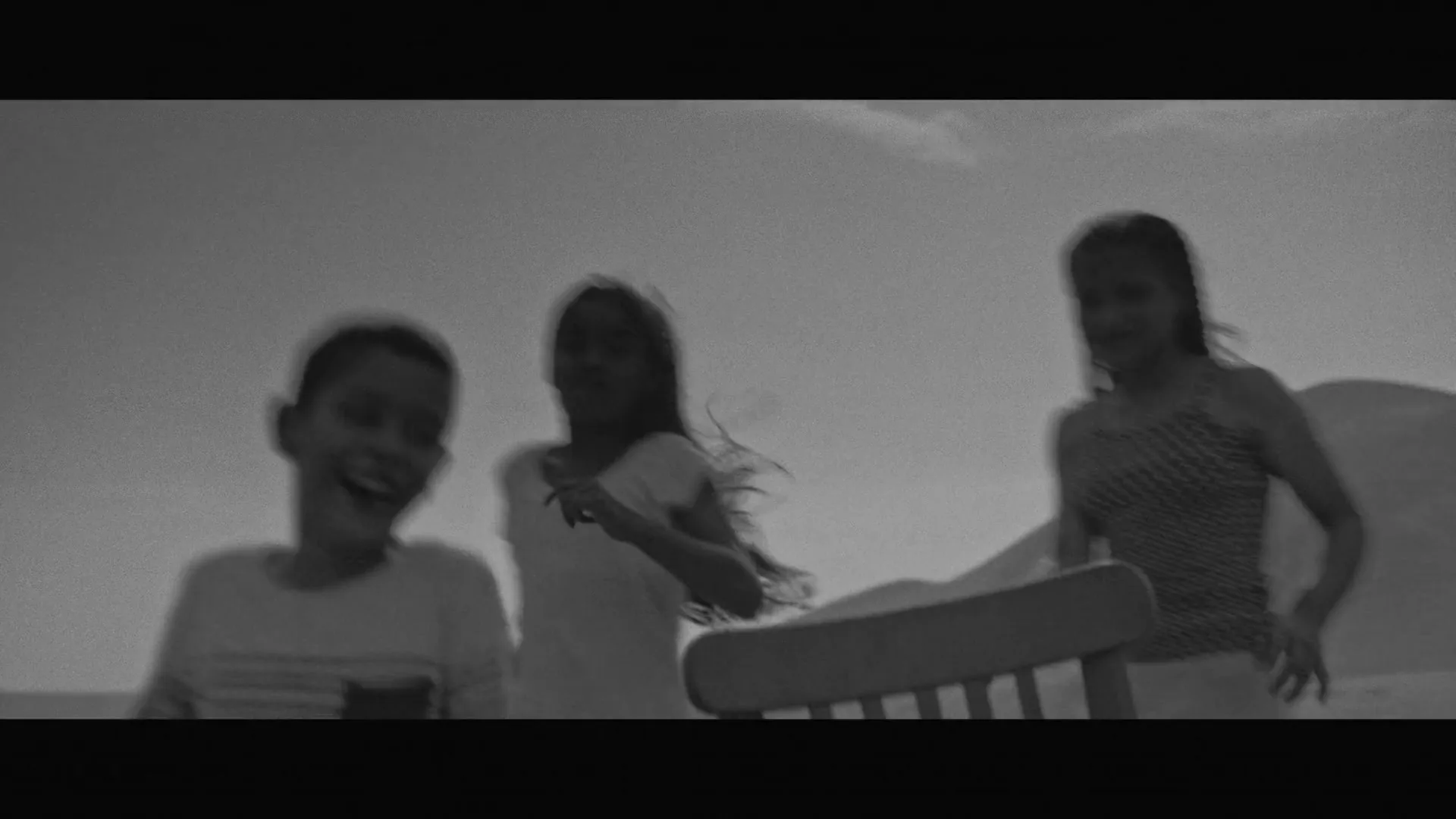 A group of children play musical chairs outdoors, running around a single chair in a black and white shot.