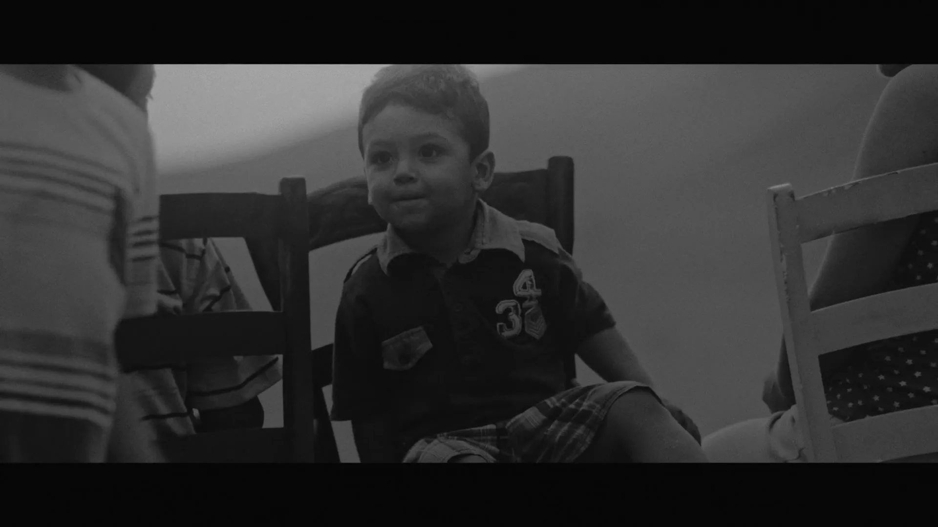 A young boy in a plaid shirt and shorts sits on a chair, looking around with a curious expression, then a young girl smiles.